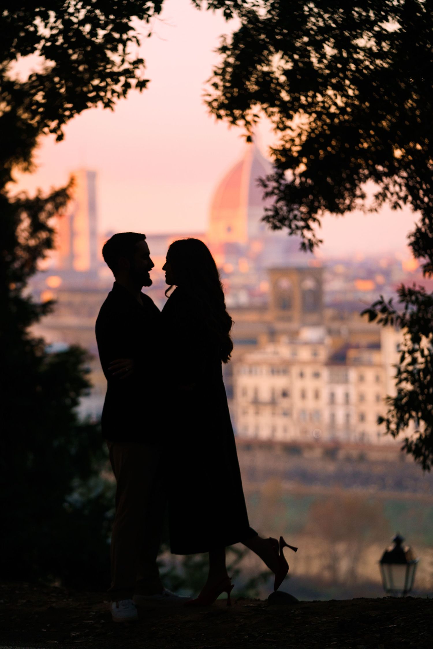 Silhouettes of two people share a romantic moment at sunset with a cityscape visible in the background.