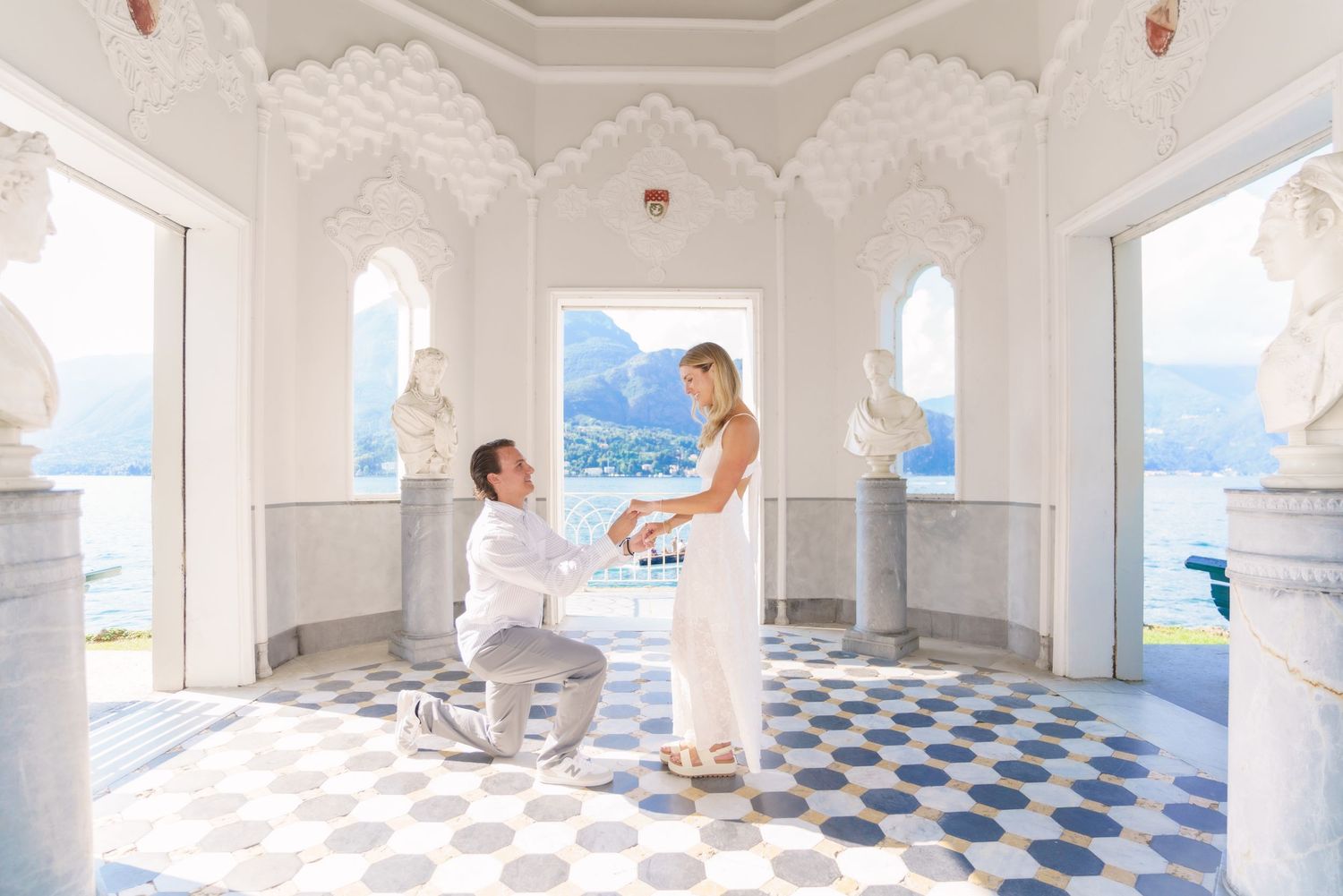 A romantic proposal moment in an elegant white pavilion overlooking a scenic lake with mountains in the background.