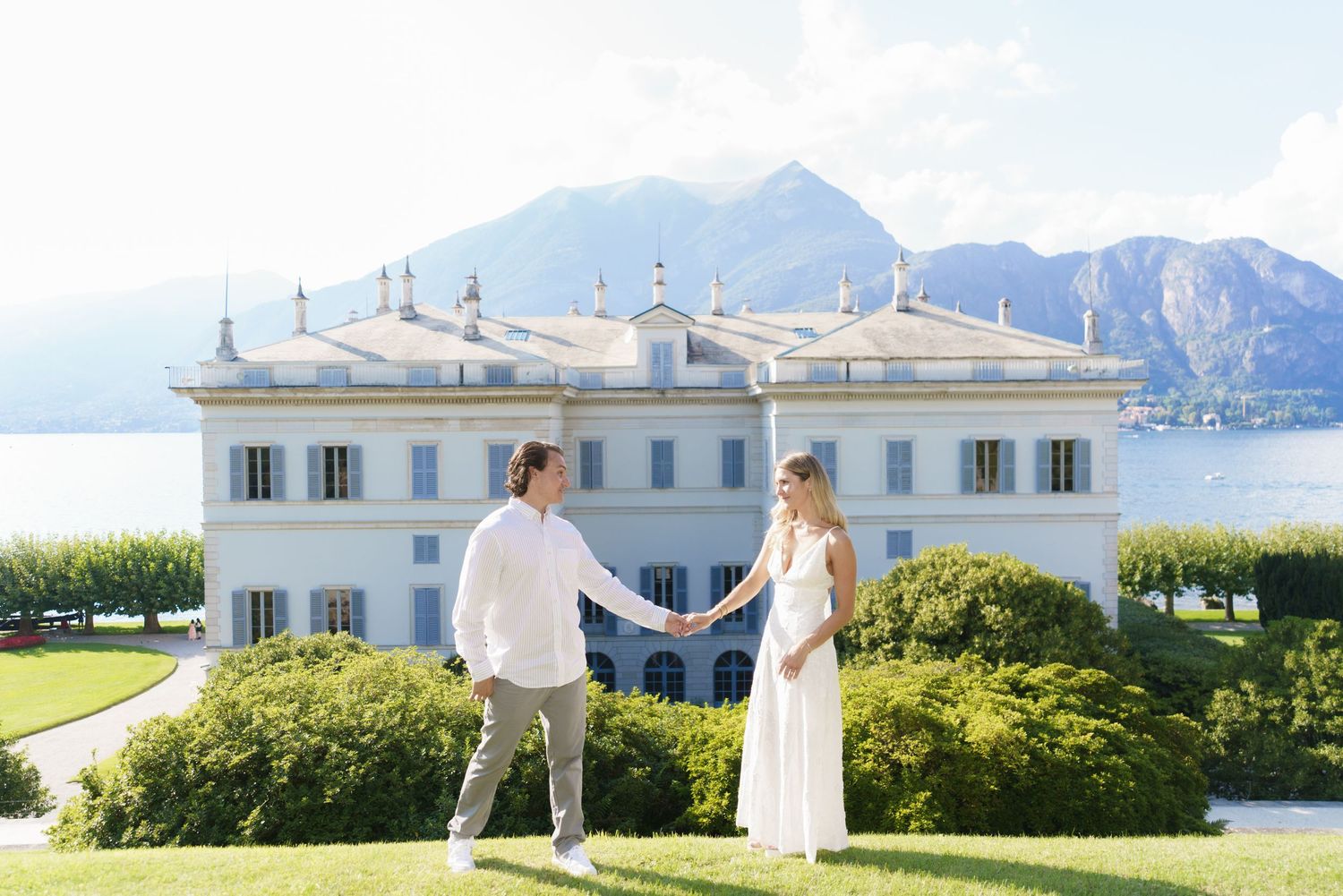 Elegant wedding couple poses at a scenic coastal venue with white buildings and mountain views in the background.