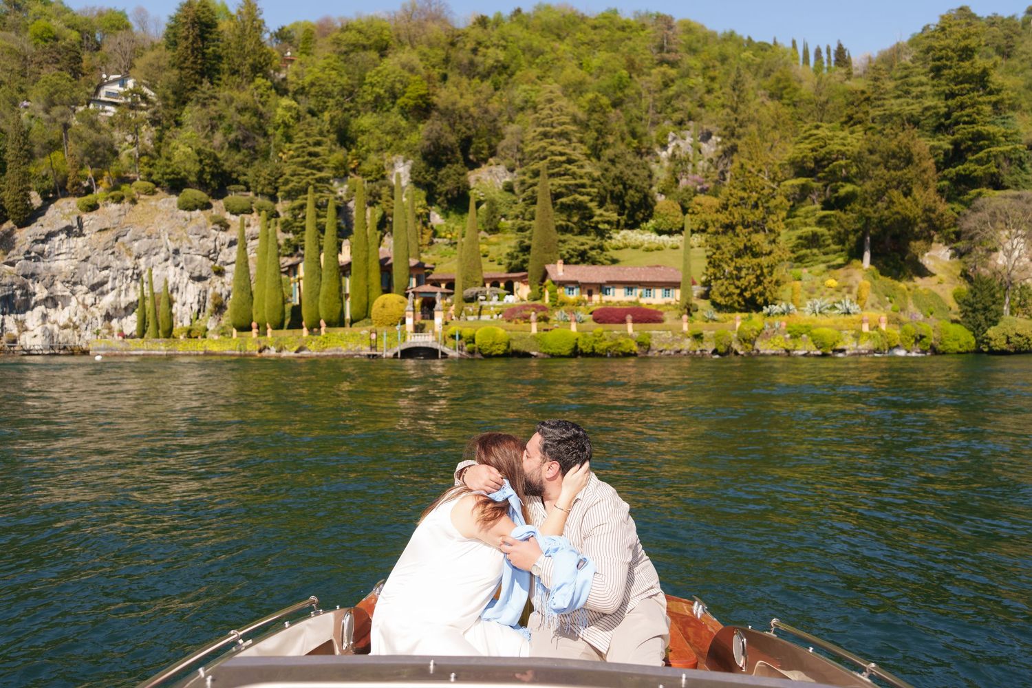 Couple kissing on a wooden boat on Lake Como with lush green hills and a villa in the background.