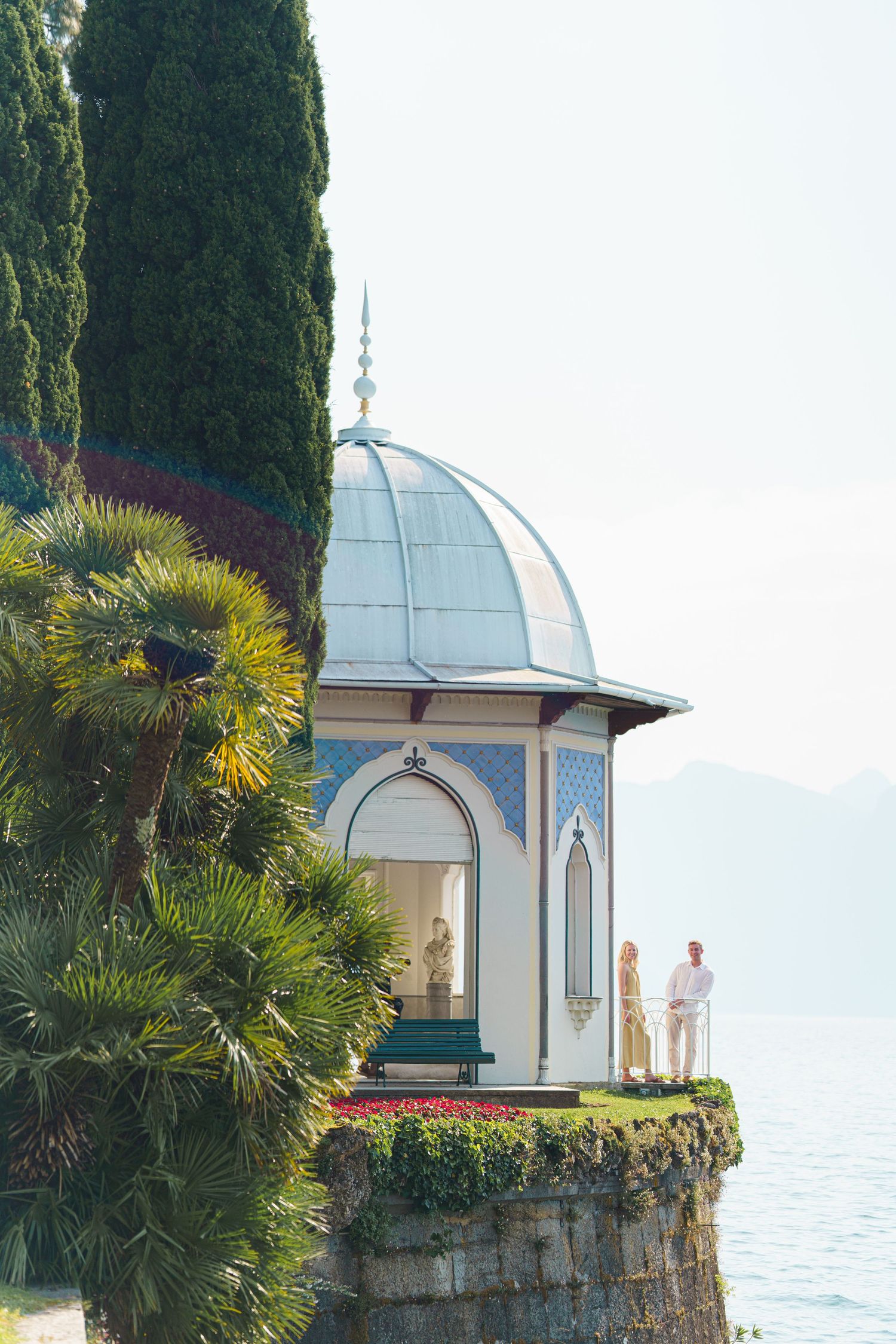 Couple standing by ornate domed pavilion overlooking serene lake, surrounded by palm trees and cypress on sunny day.