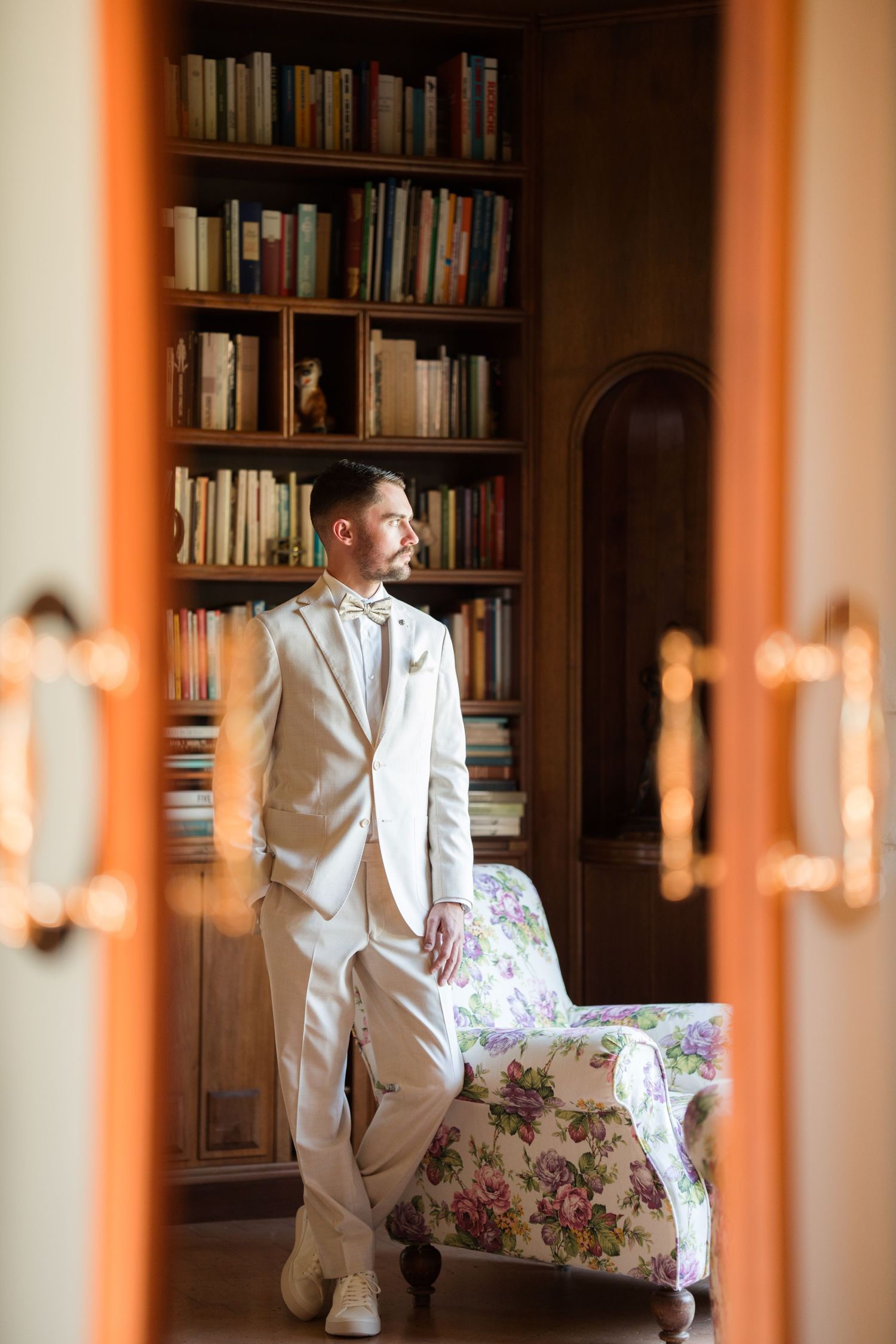 A groom in a cream suit stands beside a floral chair in an elegant library with wooden bookshelves.