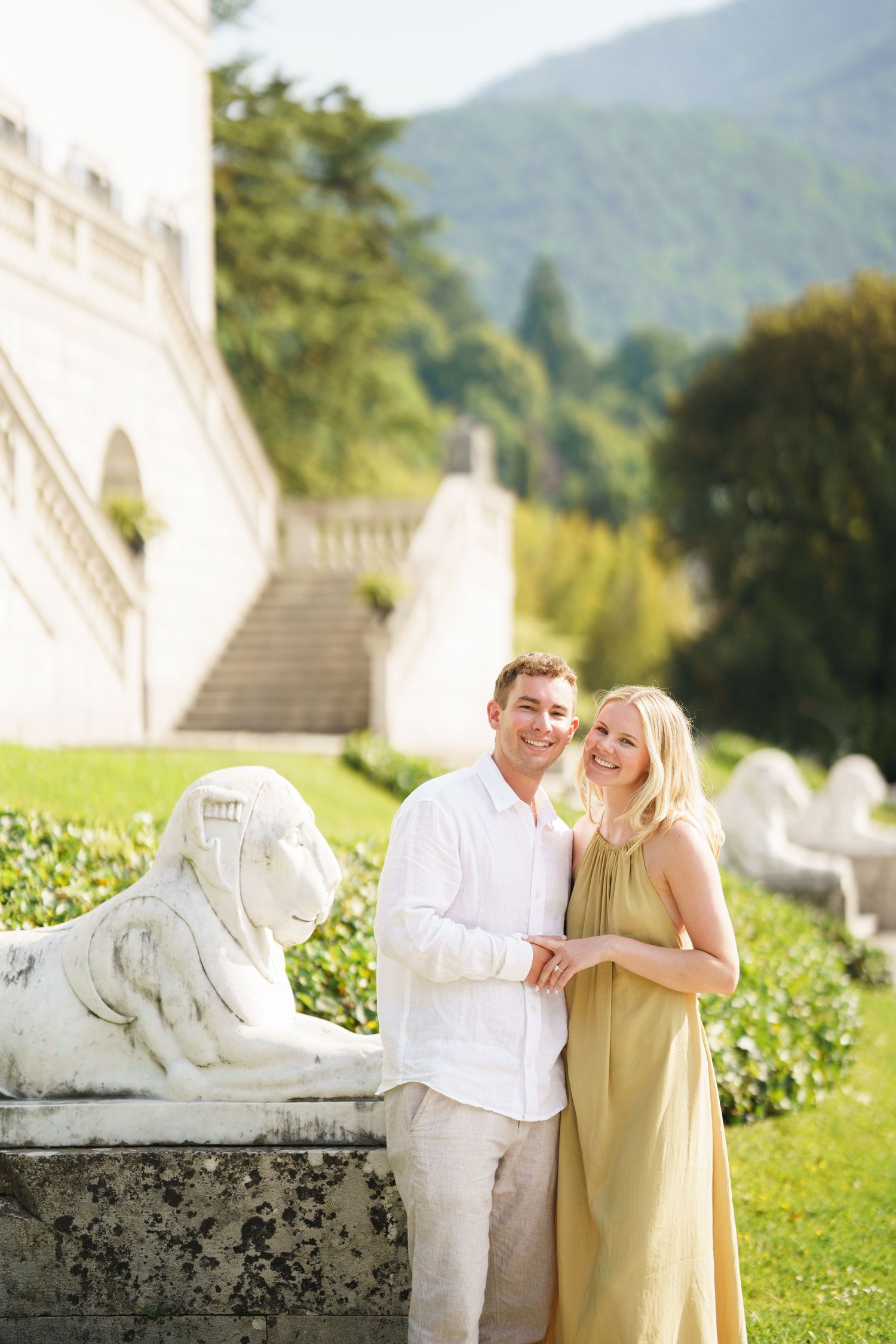 Couple posing by a stone lion sculpture near a grand estate with mountain views in the background.