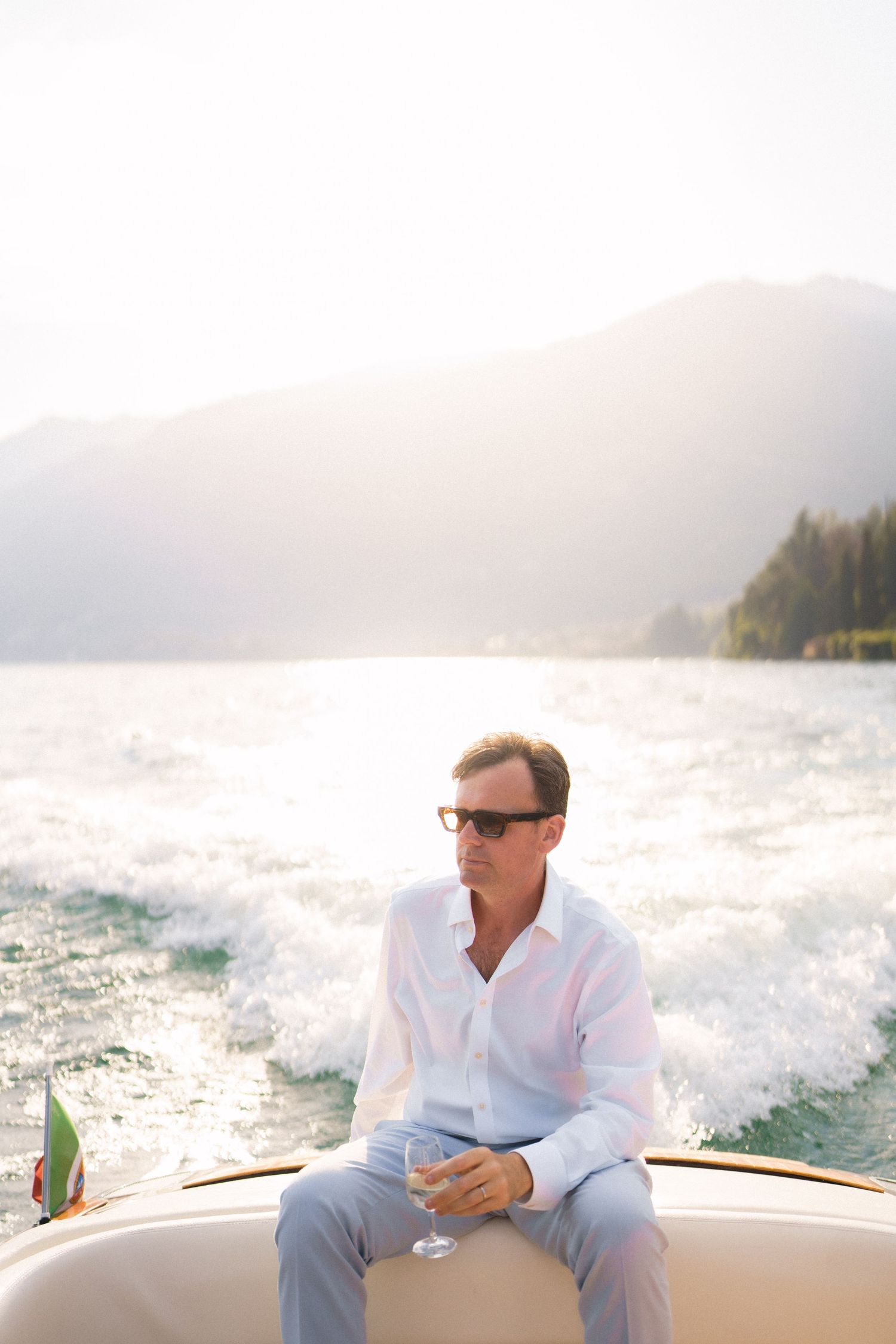 Man in white shirt and sunglasses holding champagne glass on luxury boat with mountain lake backdrop.