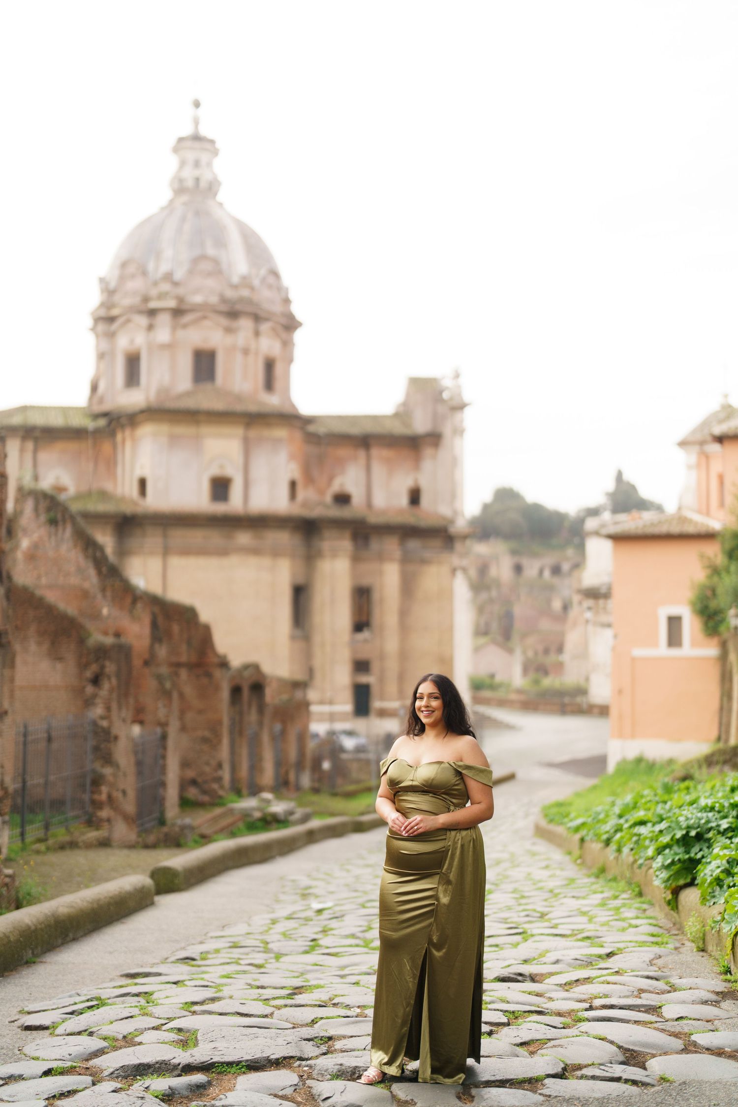 Pregnant woman in olive green gown poses on cobblestone path before ancient Roman ruins and domed church in Rome, Italy.