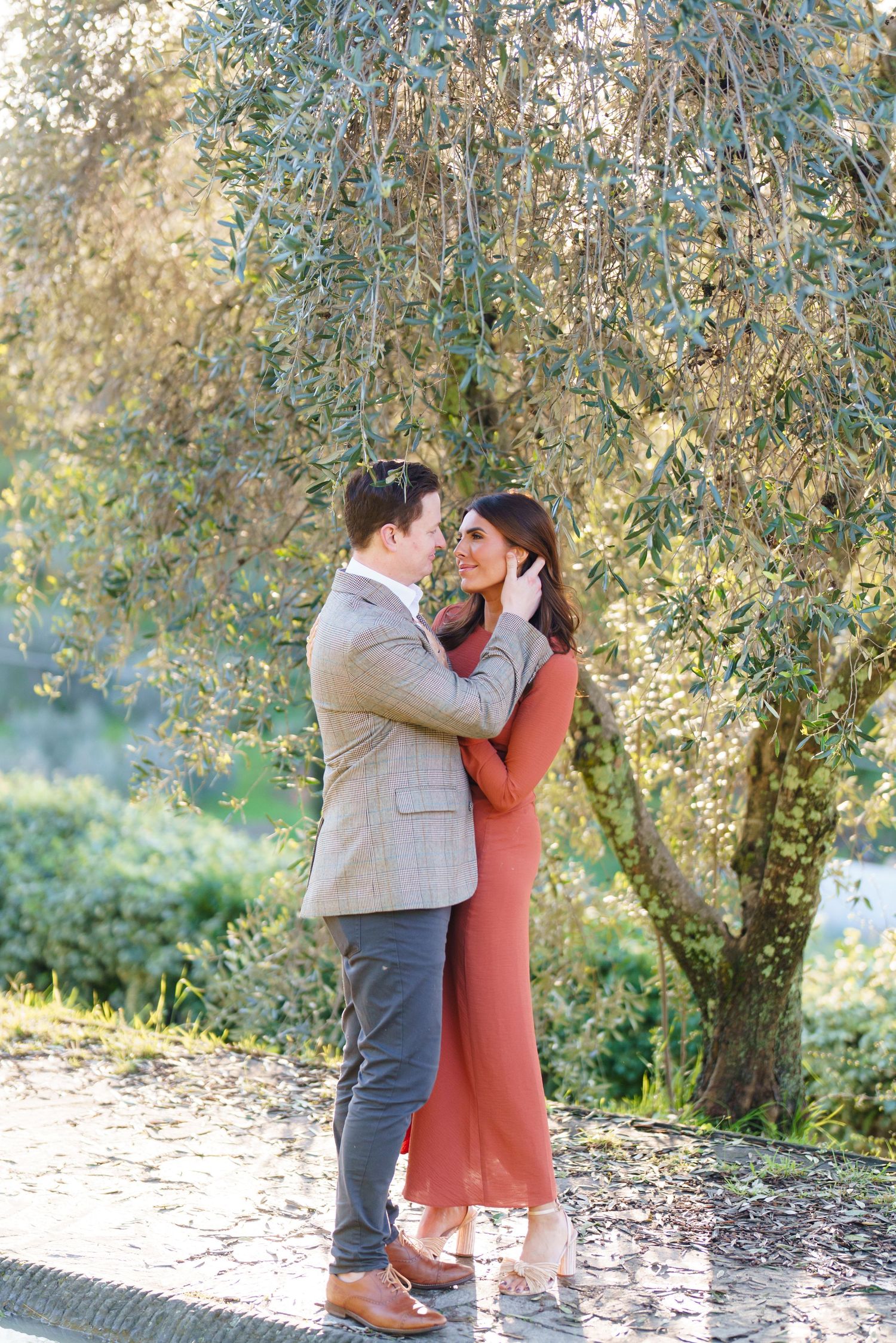 Couple embracing under an olive tree, man in tan blazer and woman in rust dress, sharing a romantic moment outdoors.