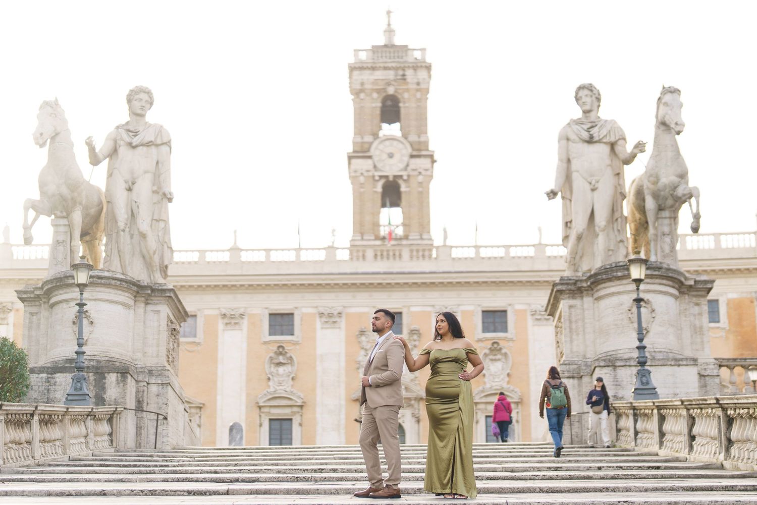 Couple posing on the steps of Piazza del Campidoglio in Rome, with classical statues and bell tower backdrop.