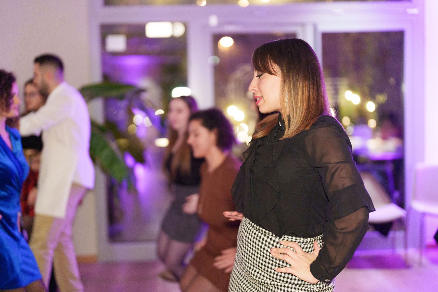 Woman in black mesh top and houndstooth skirt dancing at a party with guests in background.
