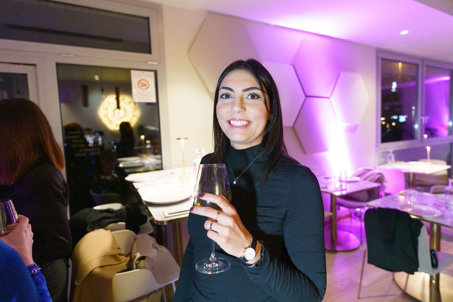 Woman in black turtleneck smiling and holding a wine glass at an elegant purple-lit restaurant event.