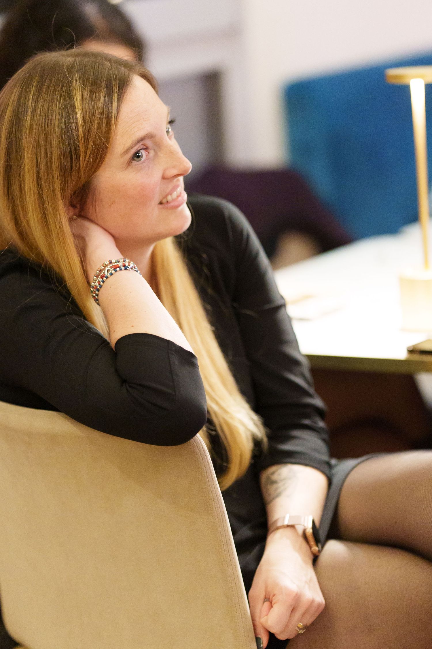 A smiling blonde woman in a black outfit sits in a chair, resting her hand on her cheek in an office setting.