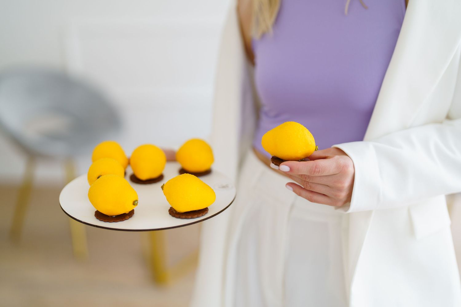 Woman in white blazer holding a plate of yellow lemon-shaped desserts and one in her hand indoors.