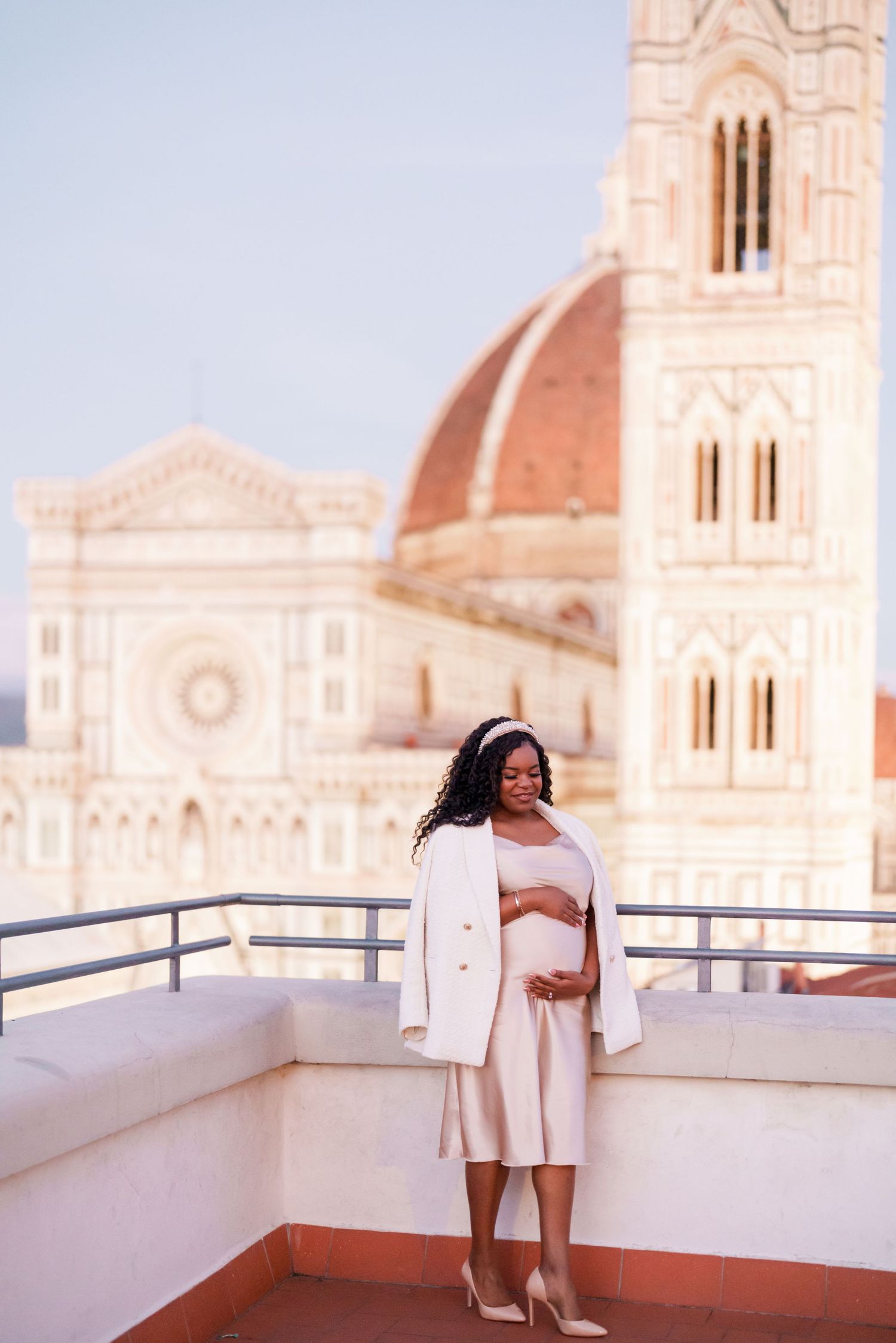 Pregnant woman in white dress and blazer on rooftop terrace with Florence Cathedral dome backdrop.