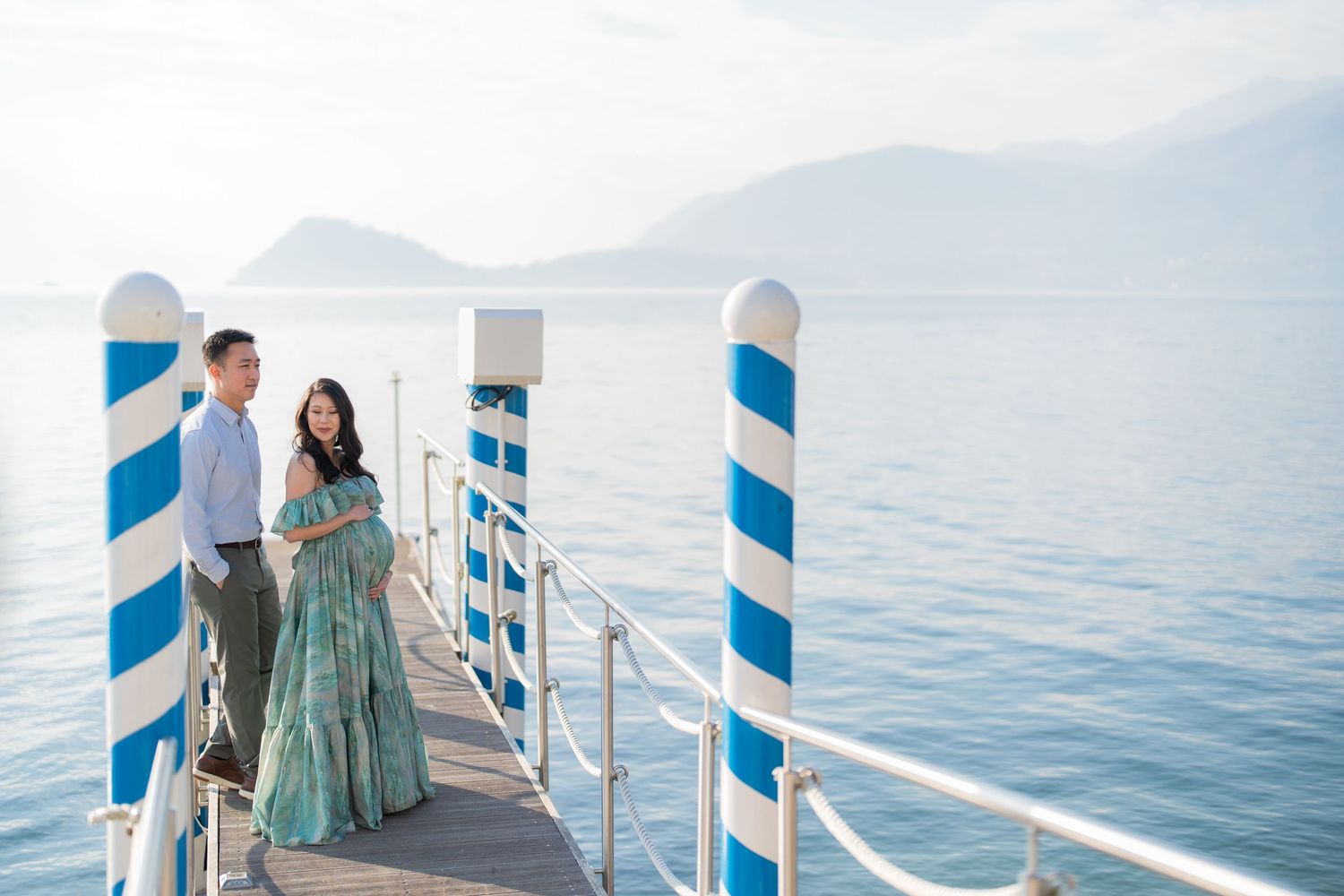 Couple standing on a white pier with blue striped posts overlooking calm blue water and mountains.