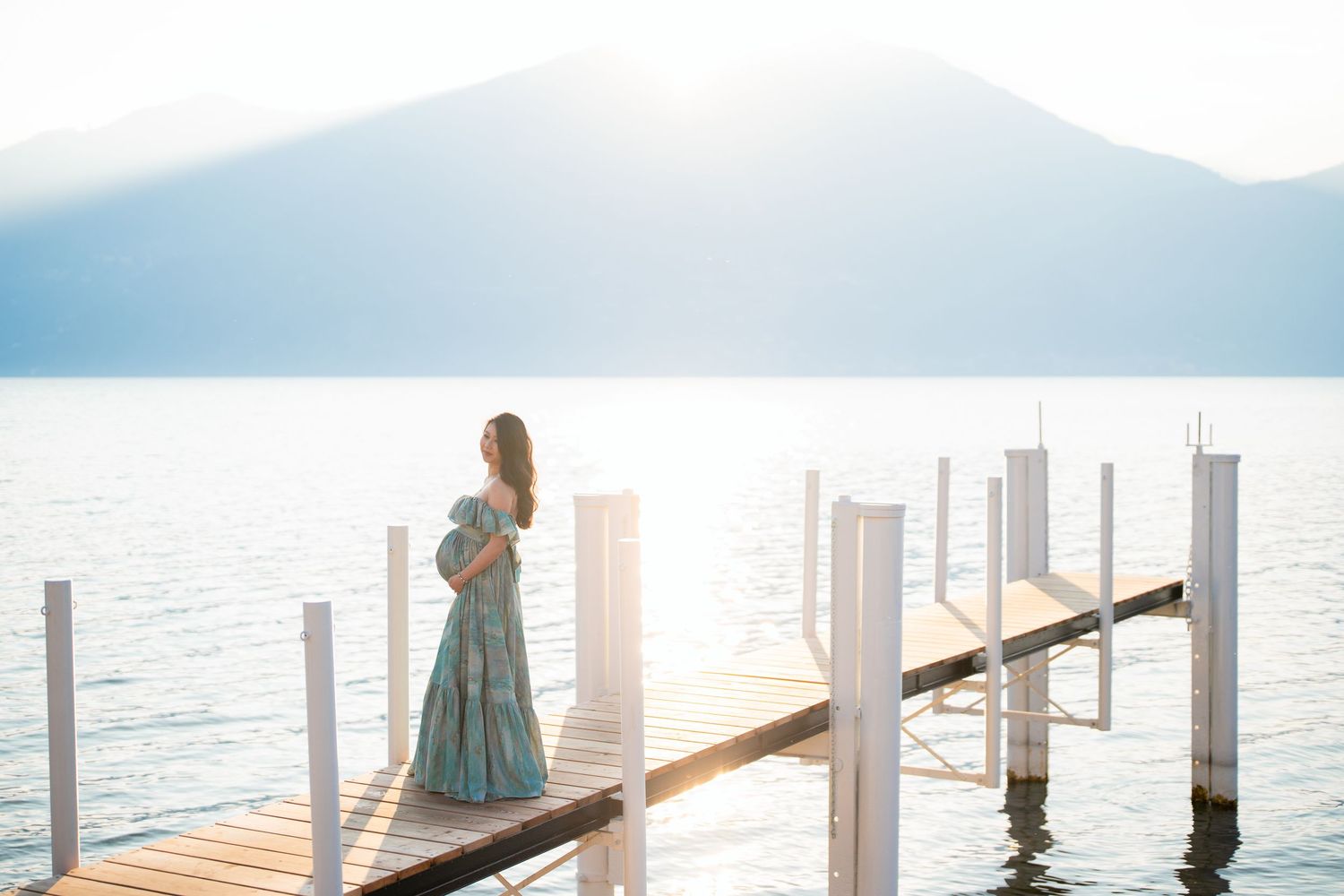 Pregnant woman in teal gown stands on wooden dock overlooking serene mountain lake at golden hour.