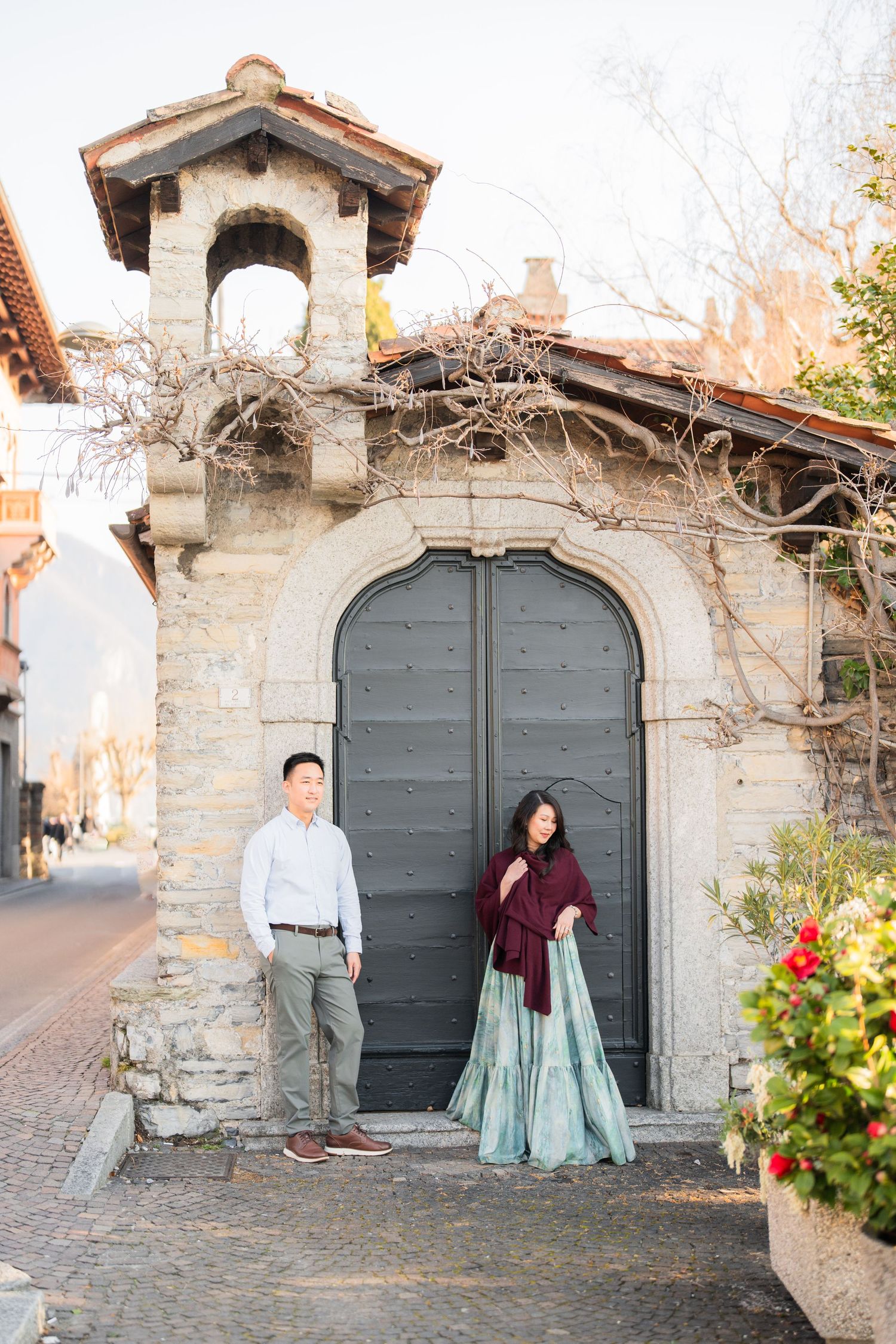 Couple posing before ornate arched stone gate in charming European village street at golden hour.