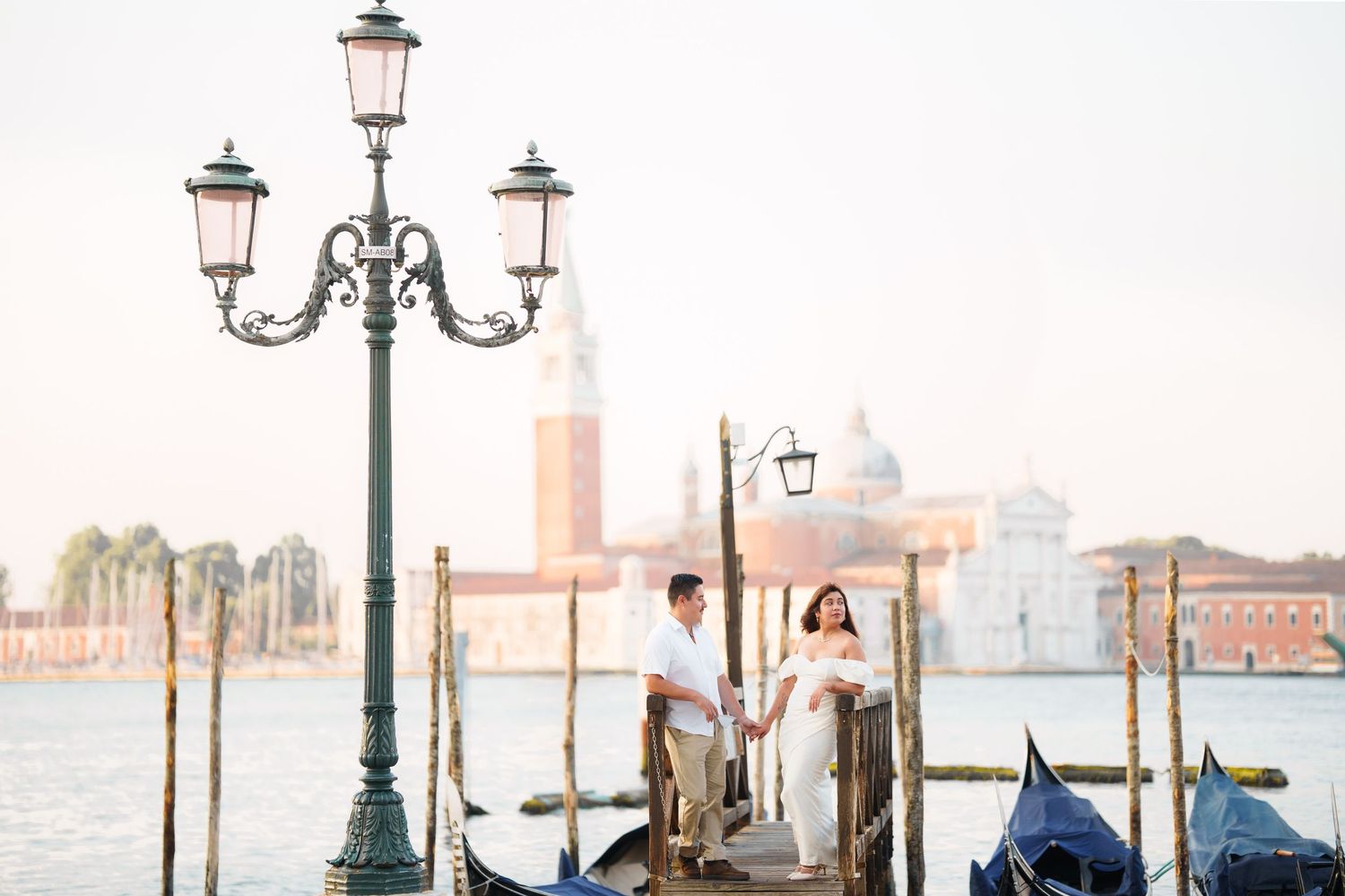 A couple stands on a dock in Venice with ornate lamp posts, gondolas, and the city skyline in soft pastel lighting.
