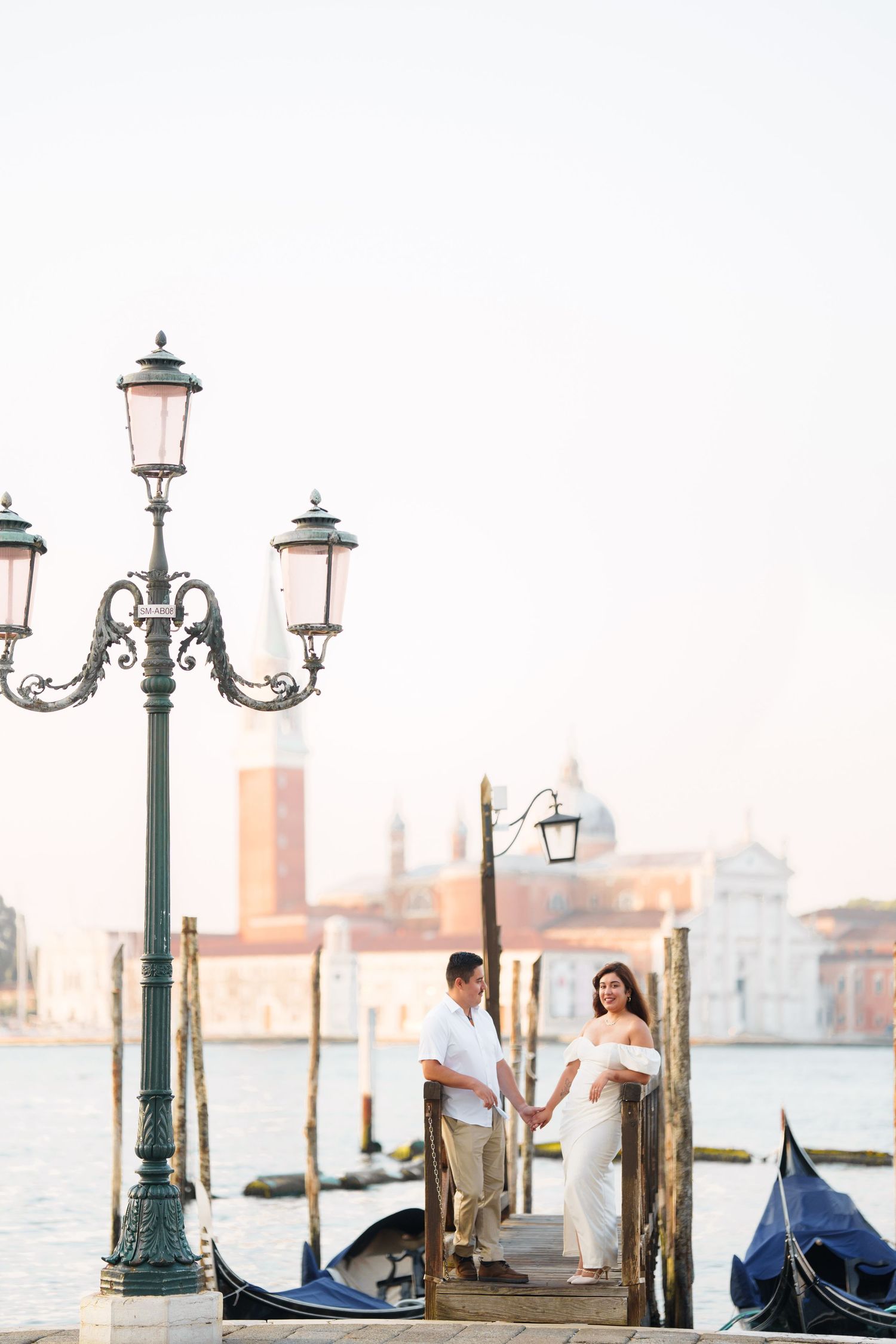 Romantic couple stands on a dock in Venice with ornate street lamp and gondolas against cityscape at sunset.
