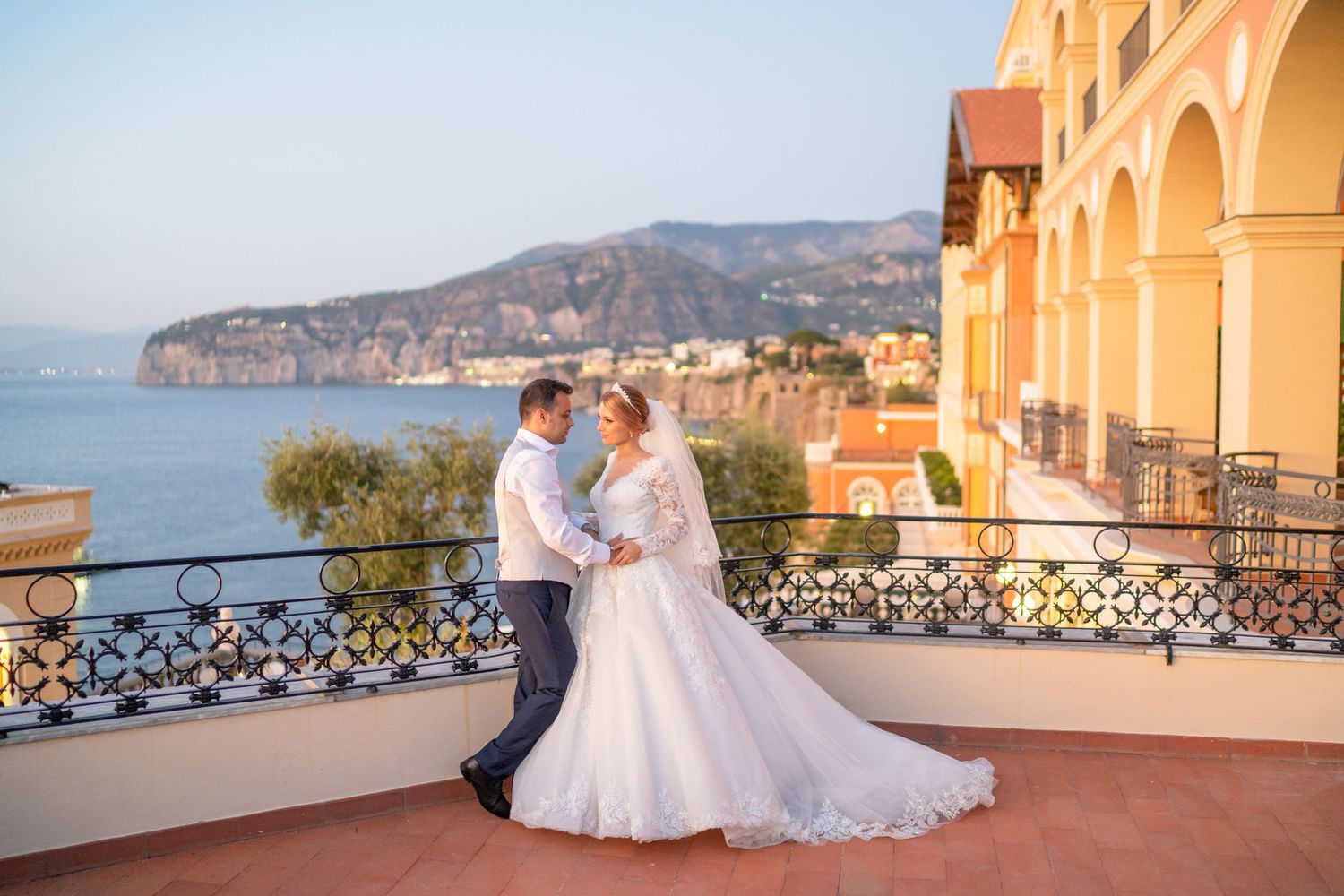 Romantic wedding couple poses on a Mediterranean terrace overlooking coastal cliffs and ocean views at sunset.
