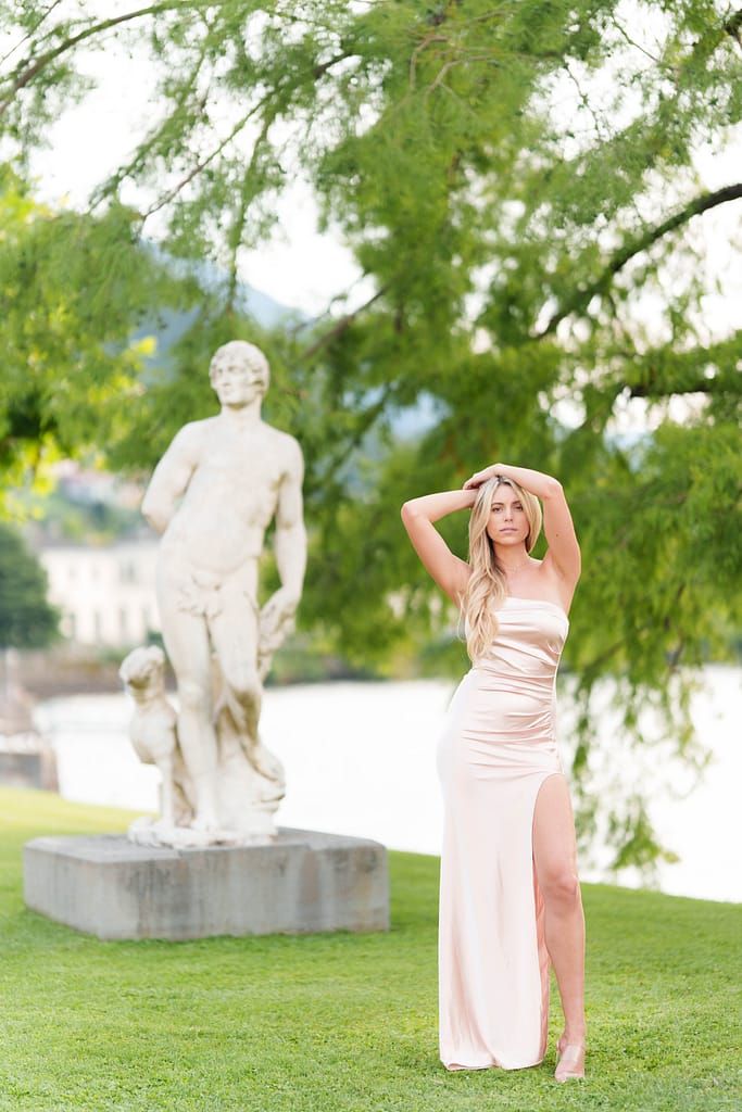 A person in a champagne-colored satin gown poses elegantly next to a classical stone statue in a garden setting.