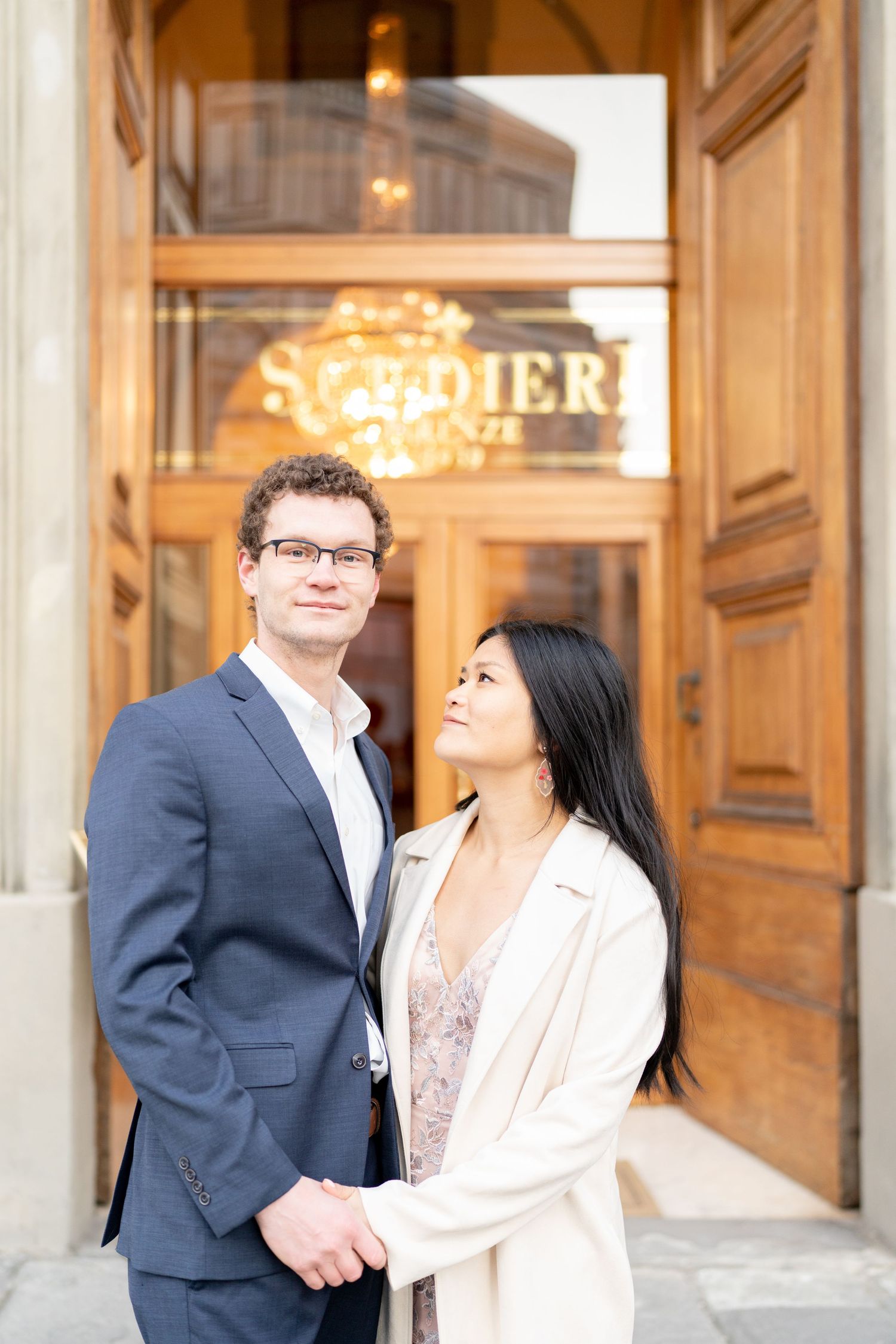 A couple shares romantic moments in front of an elegant building with ornate doors and warm lighting.