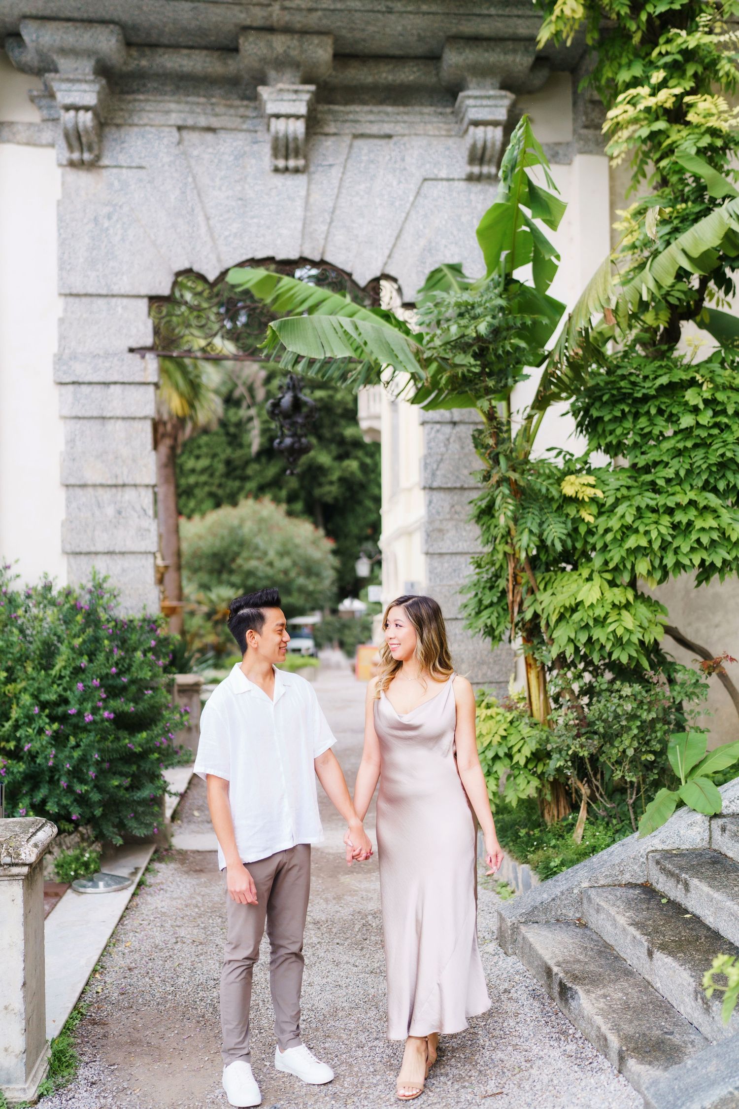 Couple walking hand in hand through an elegant stone archway surrounded by lush greenery in a romantic garden setting.