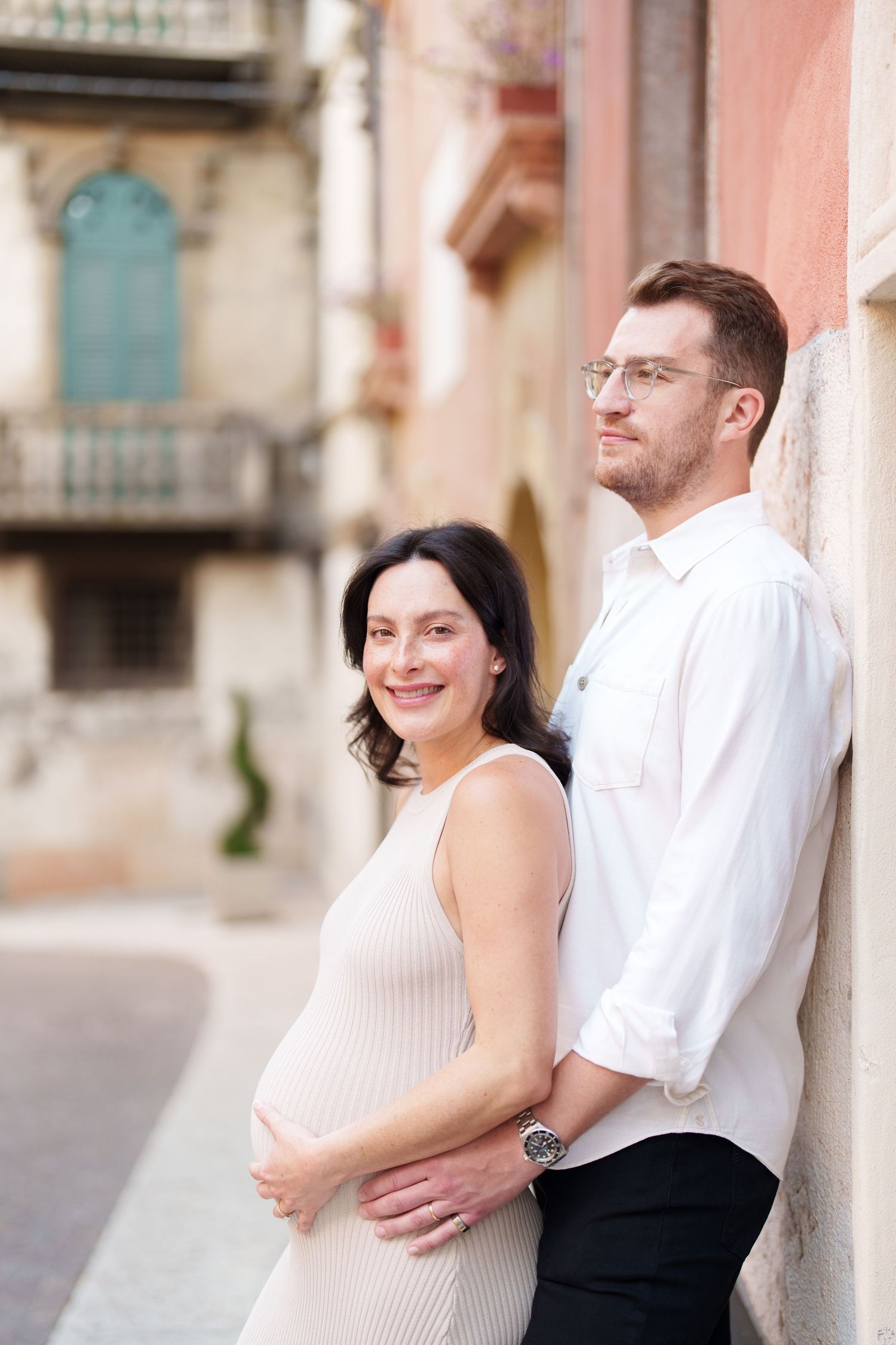 A couple in white attire poses together for romantic maternity photos on a city street.
