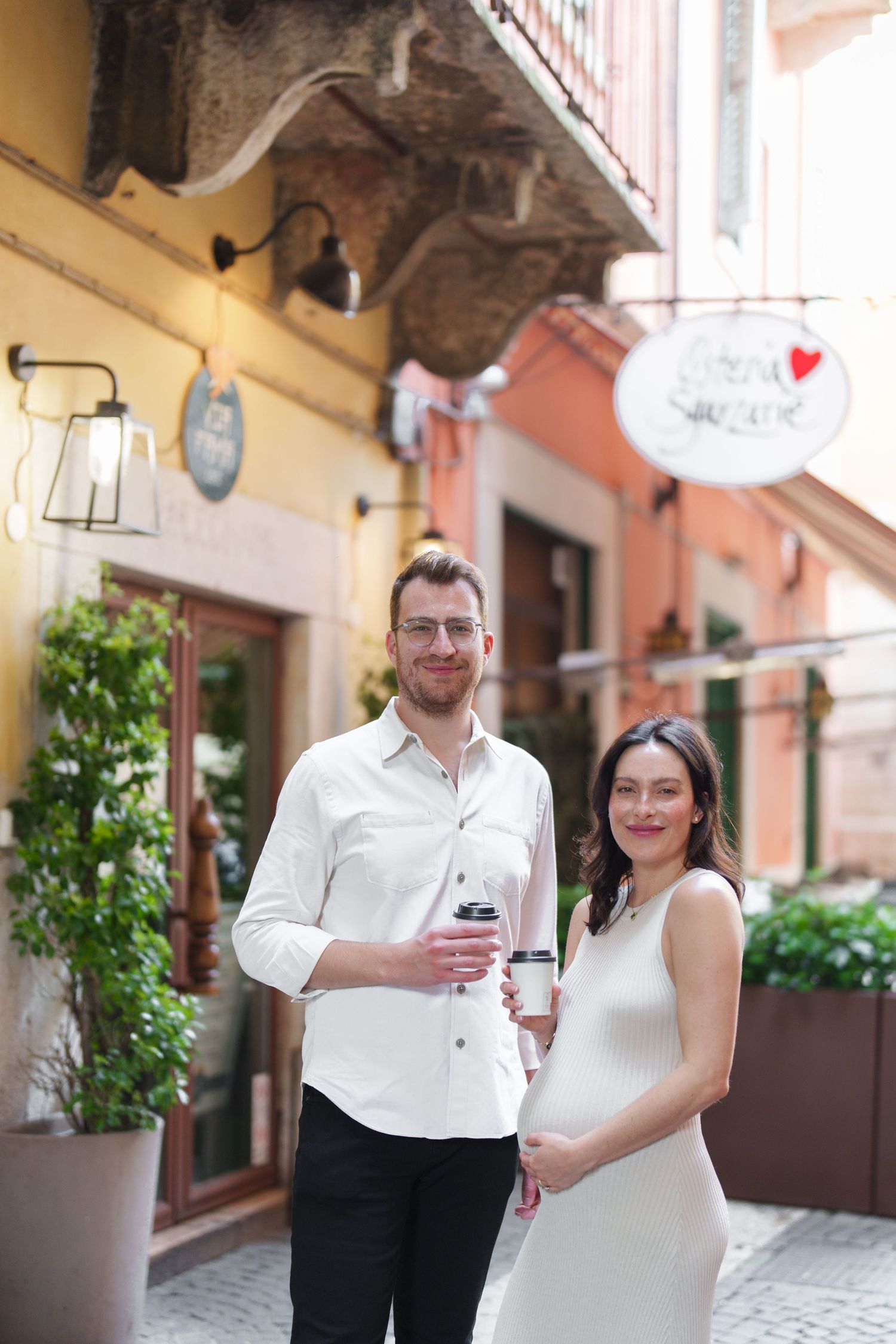 A couple in formal wedding attire poses together on a charming European cobblestone street with terracotta buildings.
