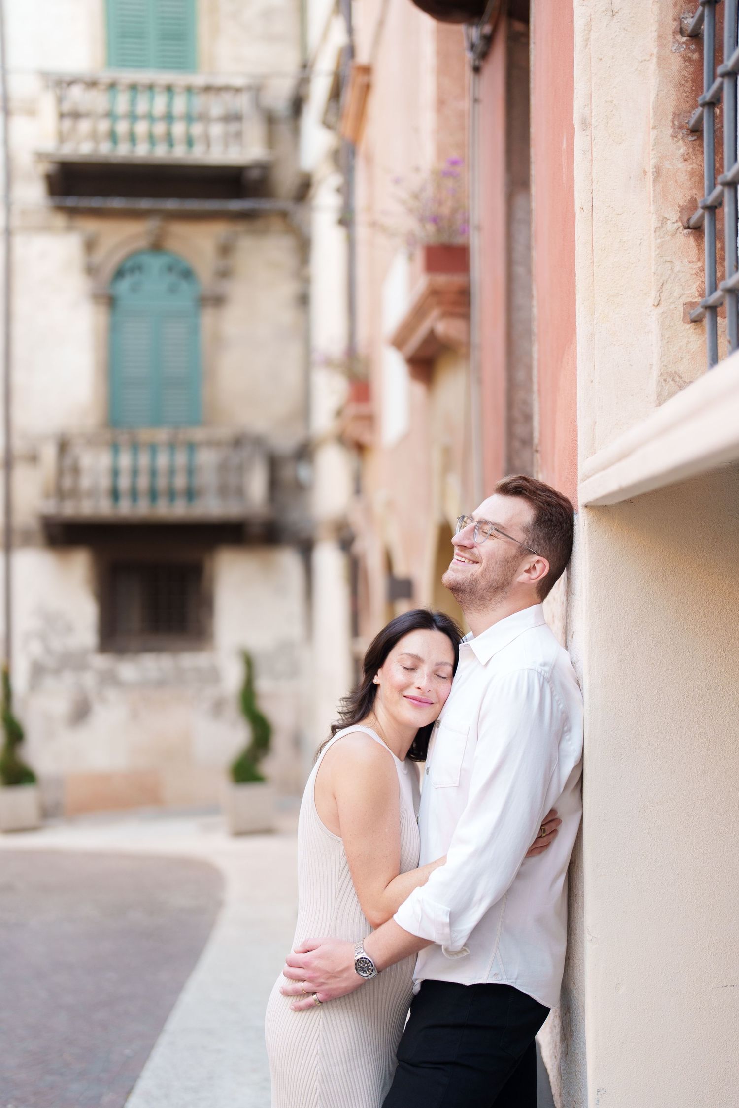 A couple embraces against a pastel wall in a European-style street with ornate balconies in the background.