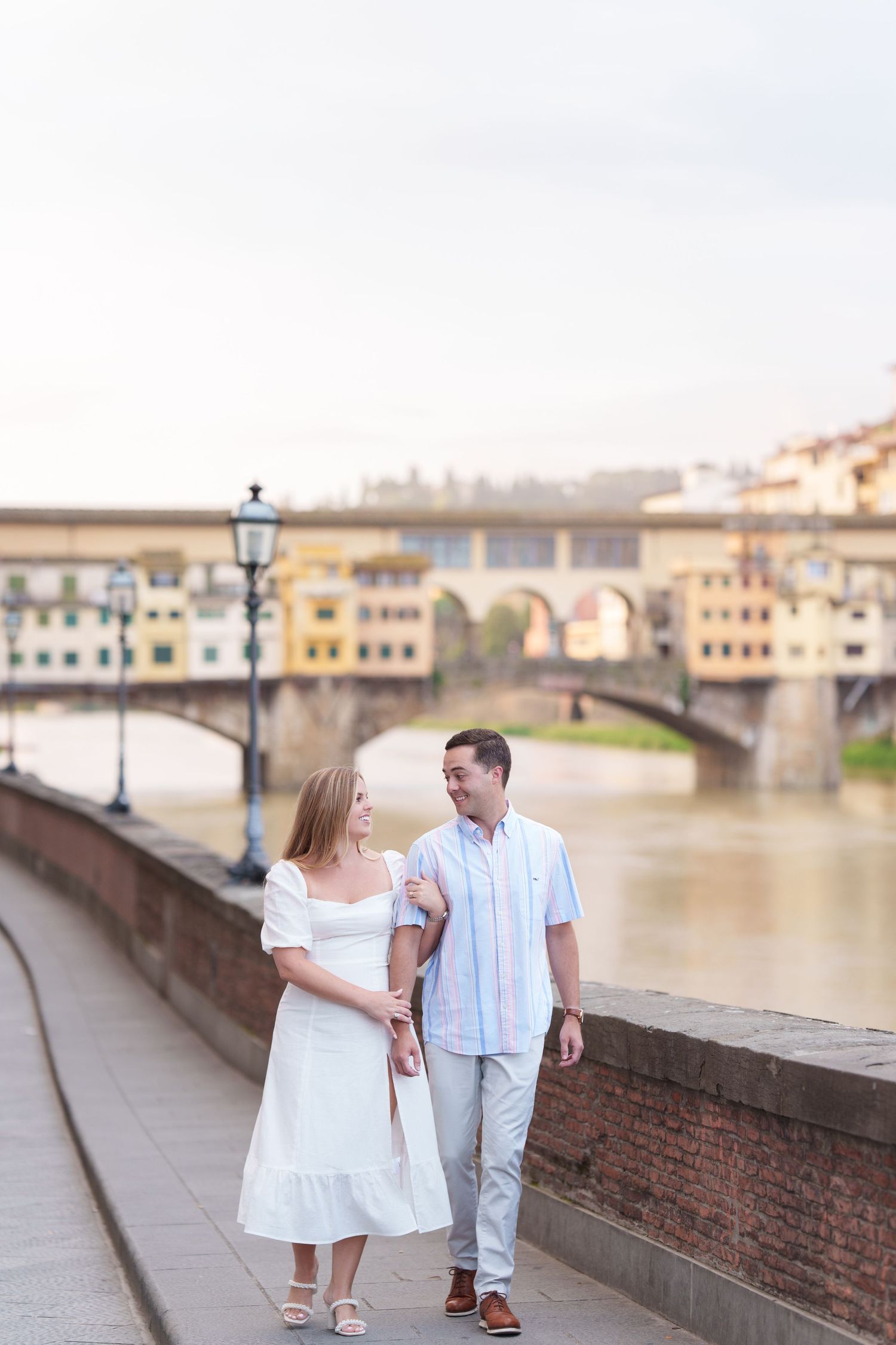 Two children in white outfits walk along a historic stone bridge overlooking a river in Florence, Italy.