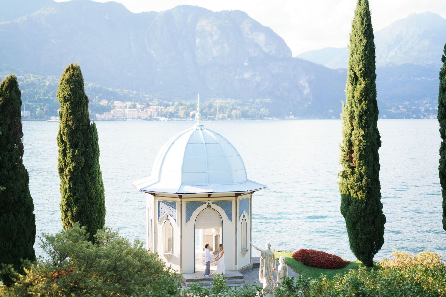 Mediterranean coastal landscape with tall cypress trees and white domed gazebo overlooking a serene lake and mountains.