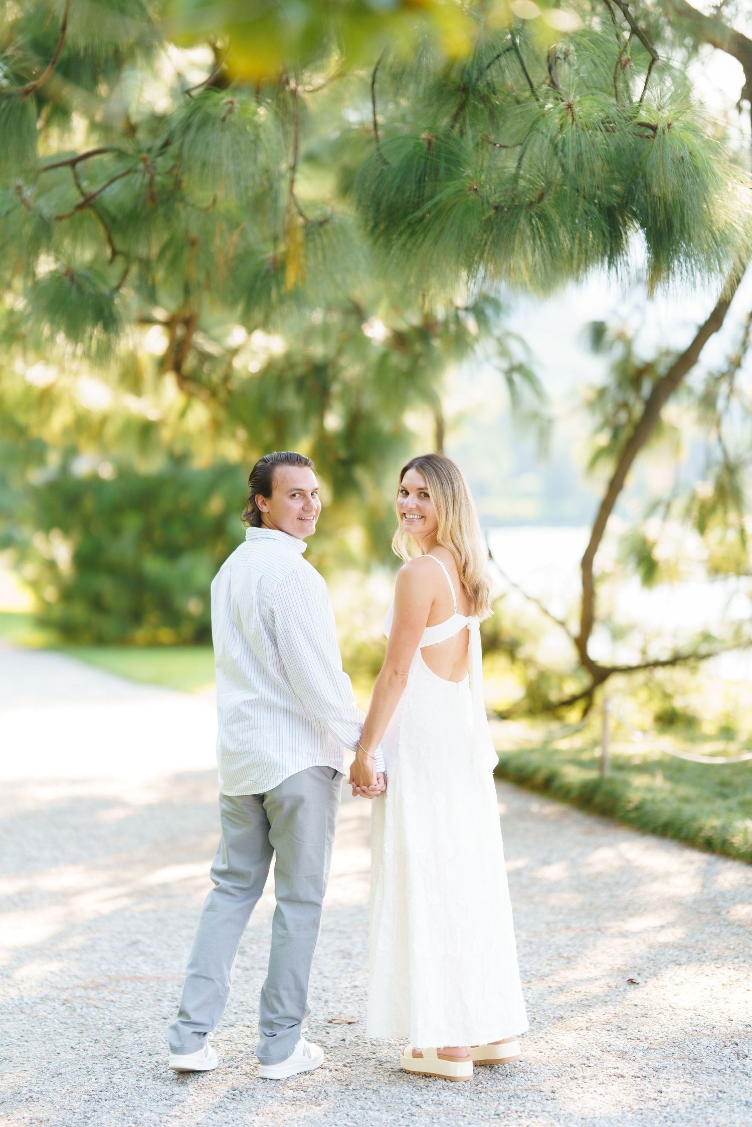 A couple in light summer attire holds hands while walking along a sunlit path under overhanging tree branches.