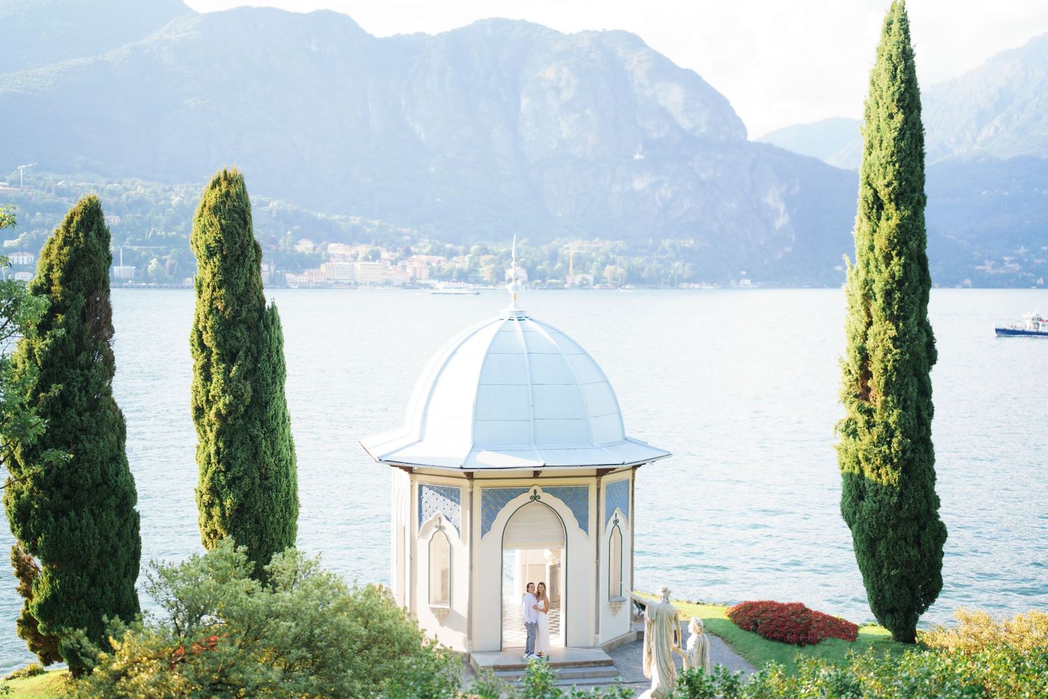A row of tall cypress trees line a scenic lakeside garden with a white gazebo overlooking mountains and water.