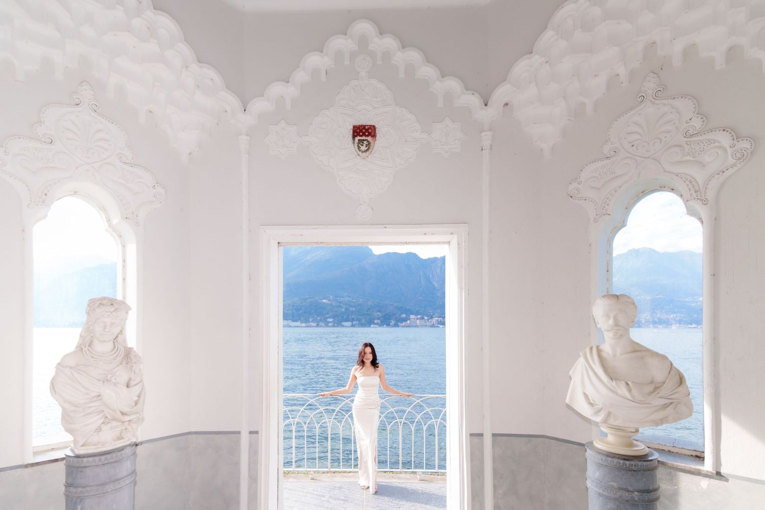 White arched colonnade overlooking a serene blue lake and mountains creates an elegant Mediterranean atmosphere.