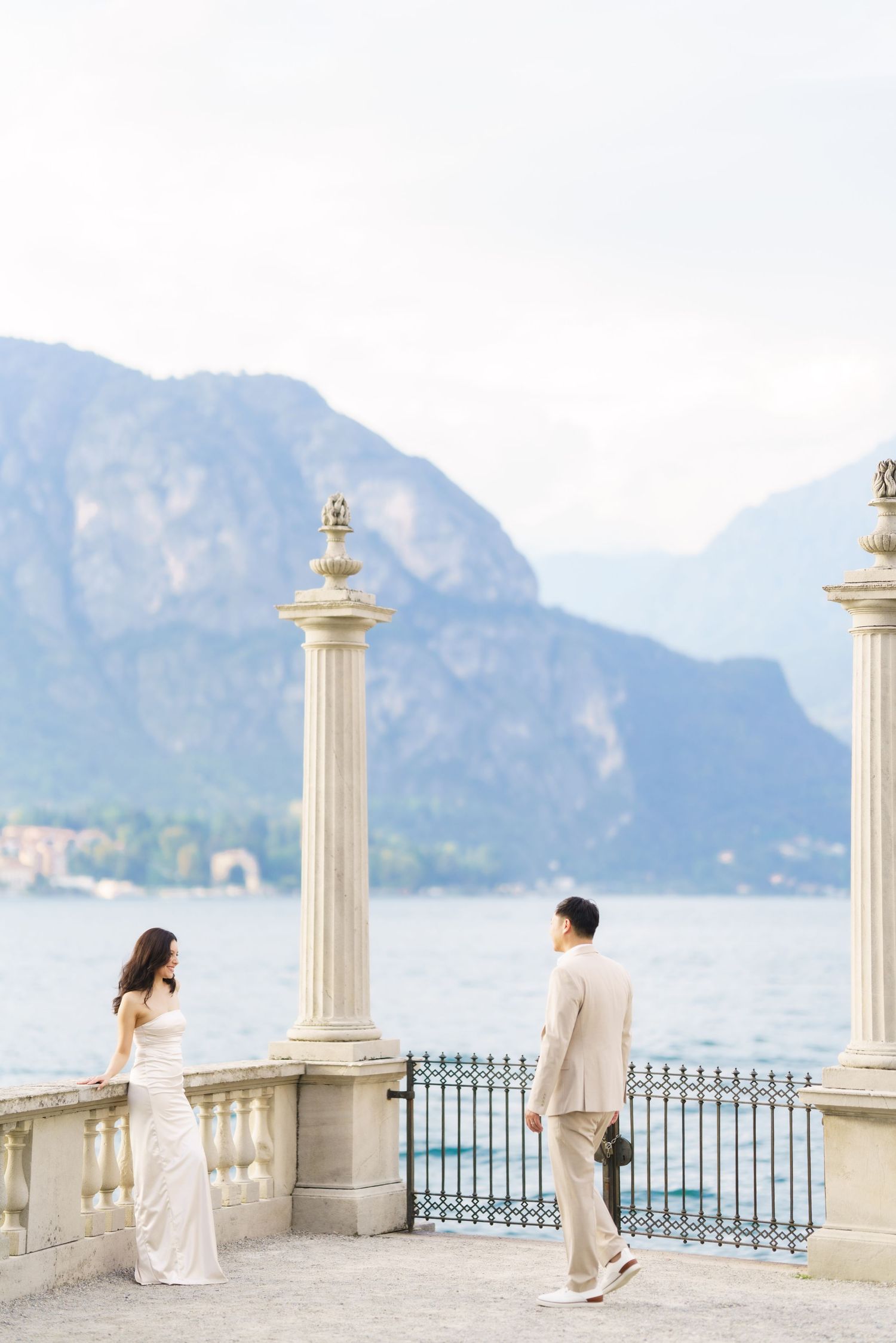 Couple in elegant attire standing on a lakeside terrace with ornate columns and mountain views in the background.