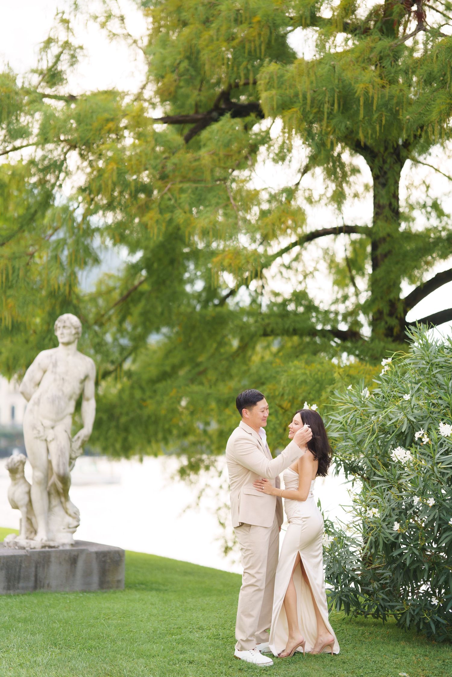 A romantic couple shares tender moments in a garden setting with classical statues and lush greenery in the background.