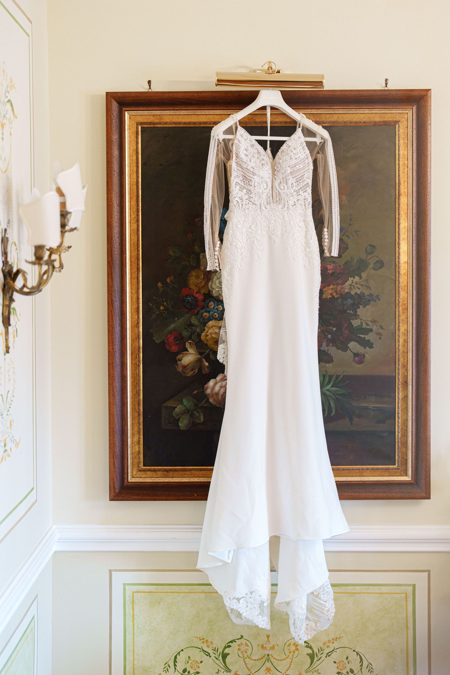 White wedding dress hanging on display against cream walls with ornate gold-framed paintings.