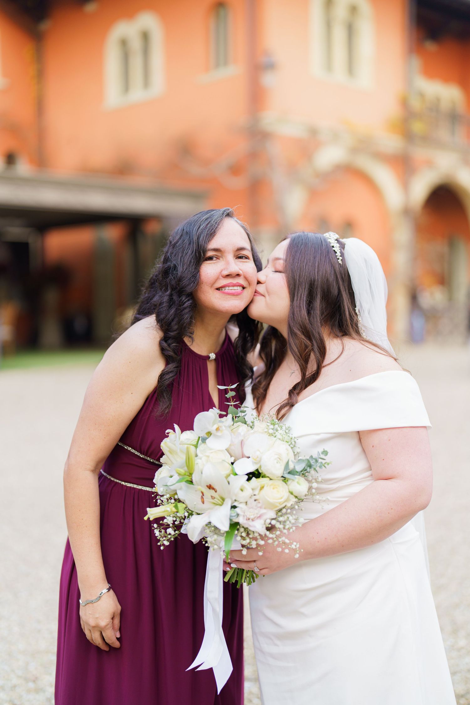 Two people embrace while wearing burgundy and white dresses at an outdoor wedding venue.