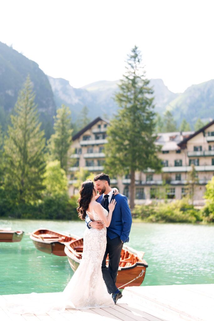 Couple shares romantic moment on wooden dock with mountain lake and boats in background.