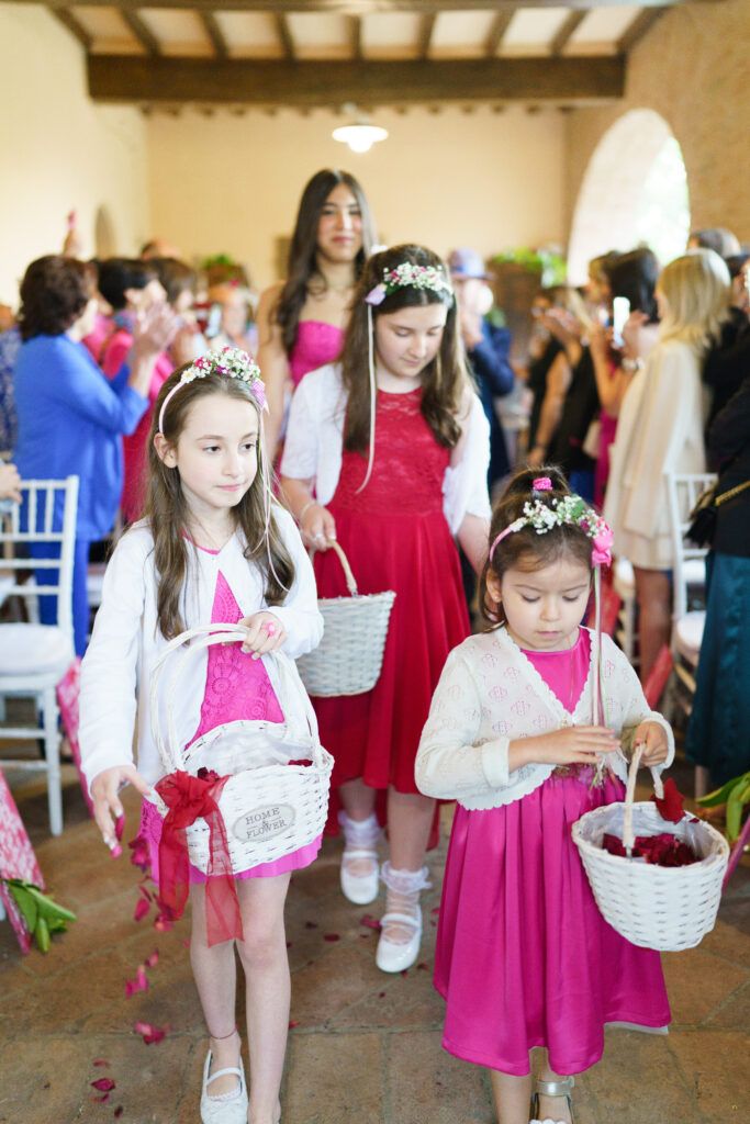 Flower girls in bright pink dresses carrying white baskets walk down the aisle.