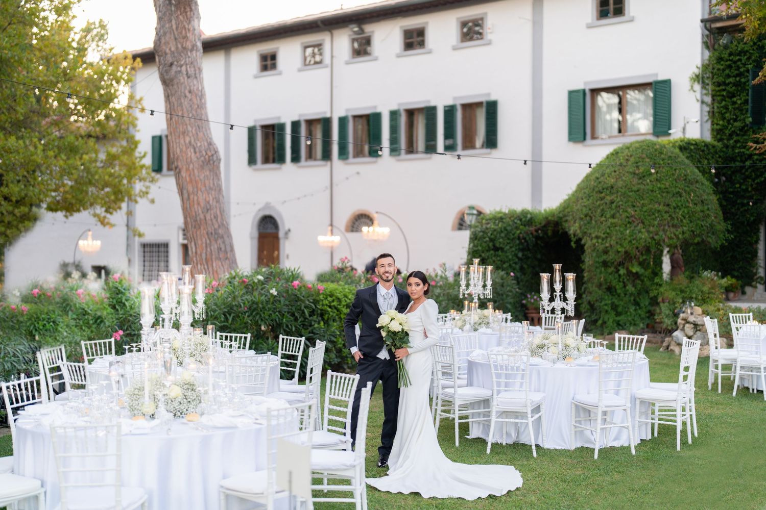 White wedding reception tables arranged on a lawn beside an Italian-style villa.