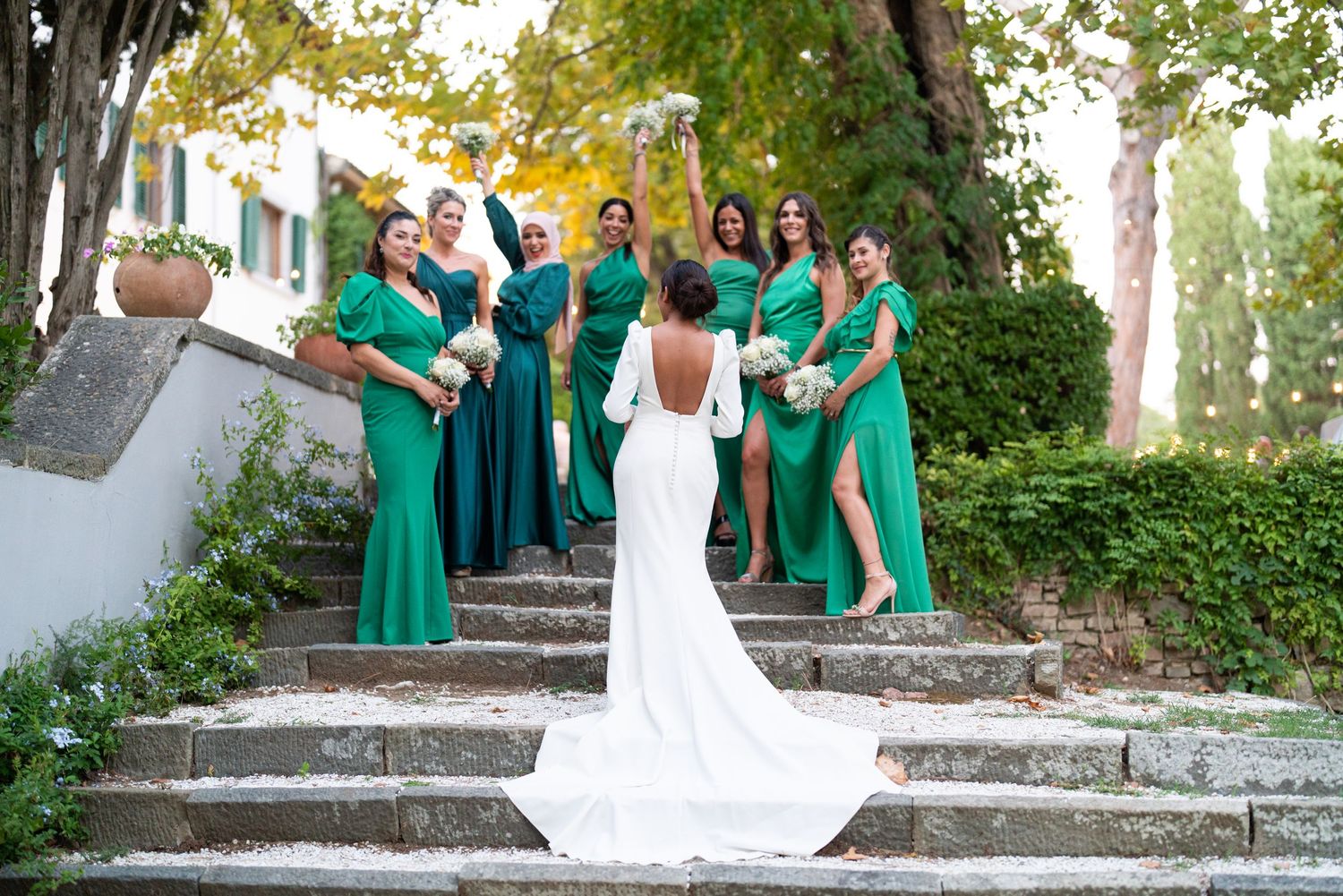 Wedding party in emerald green dresses posing on stone steps in a garden setting.