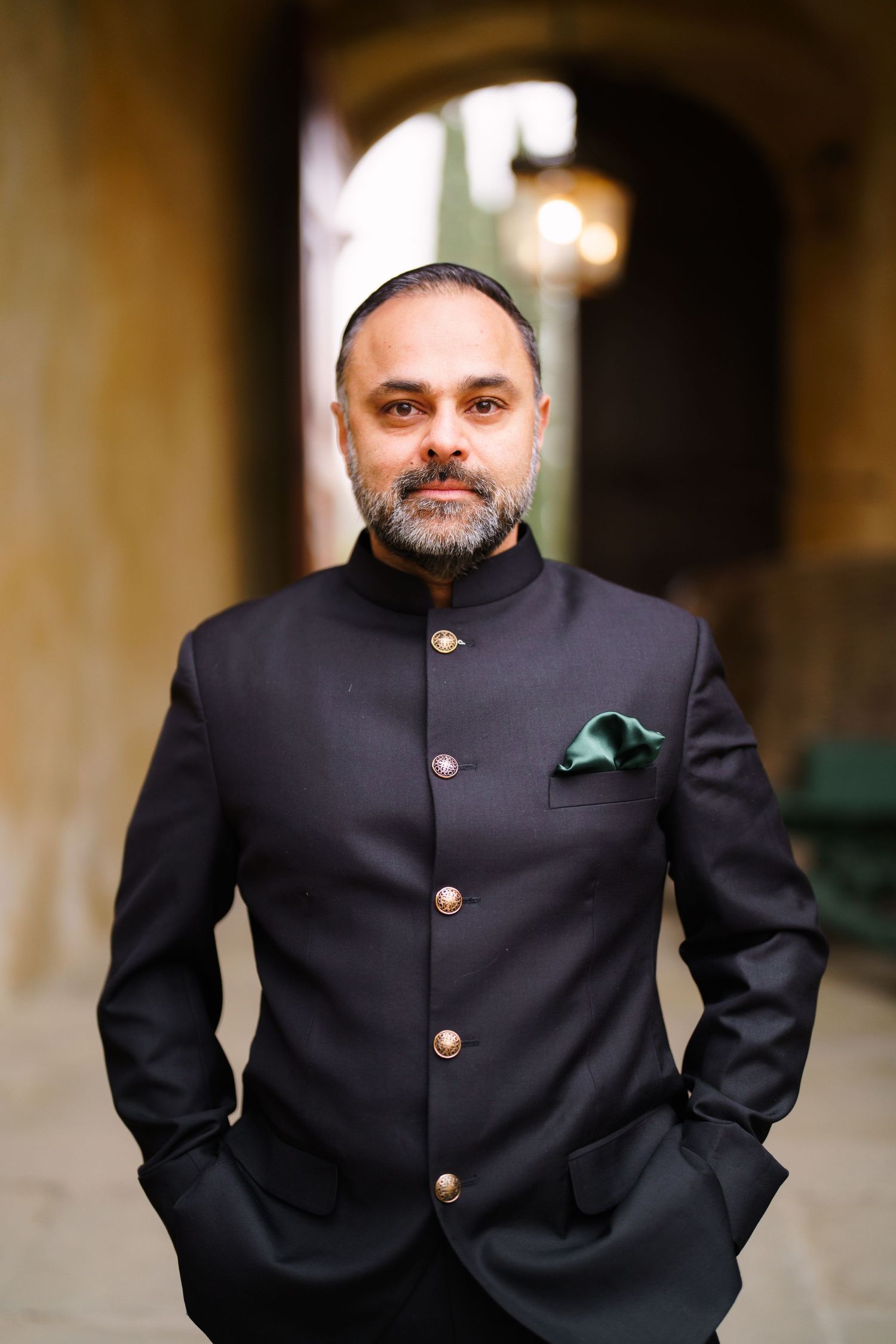 Portrait of person wearing a formal black Nehru-style jacket with brass buttons against a blurred background.