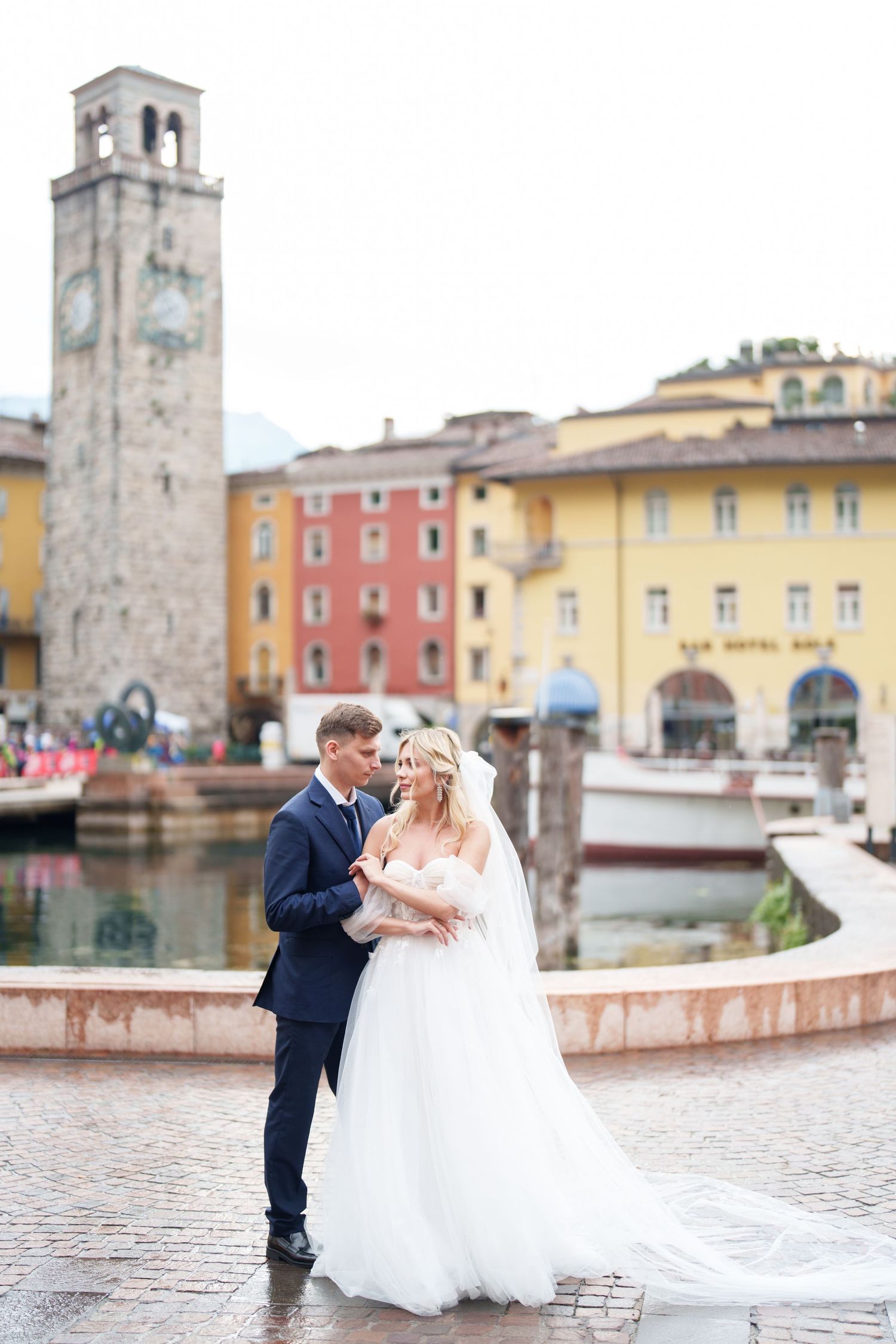 A romantic wedding couple stands by a historic clock tower and colorful buildings along a harbor in Riva del Garda, Italy.