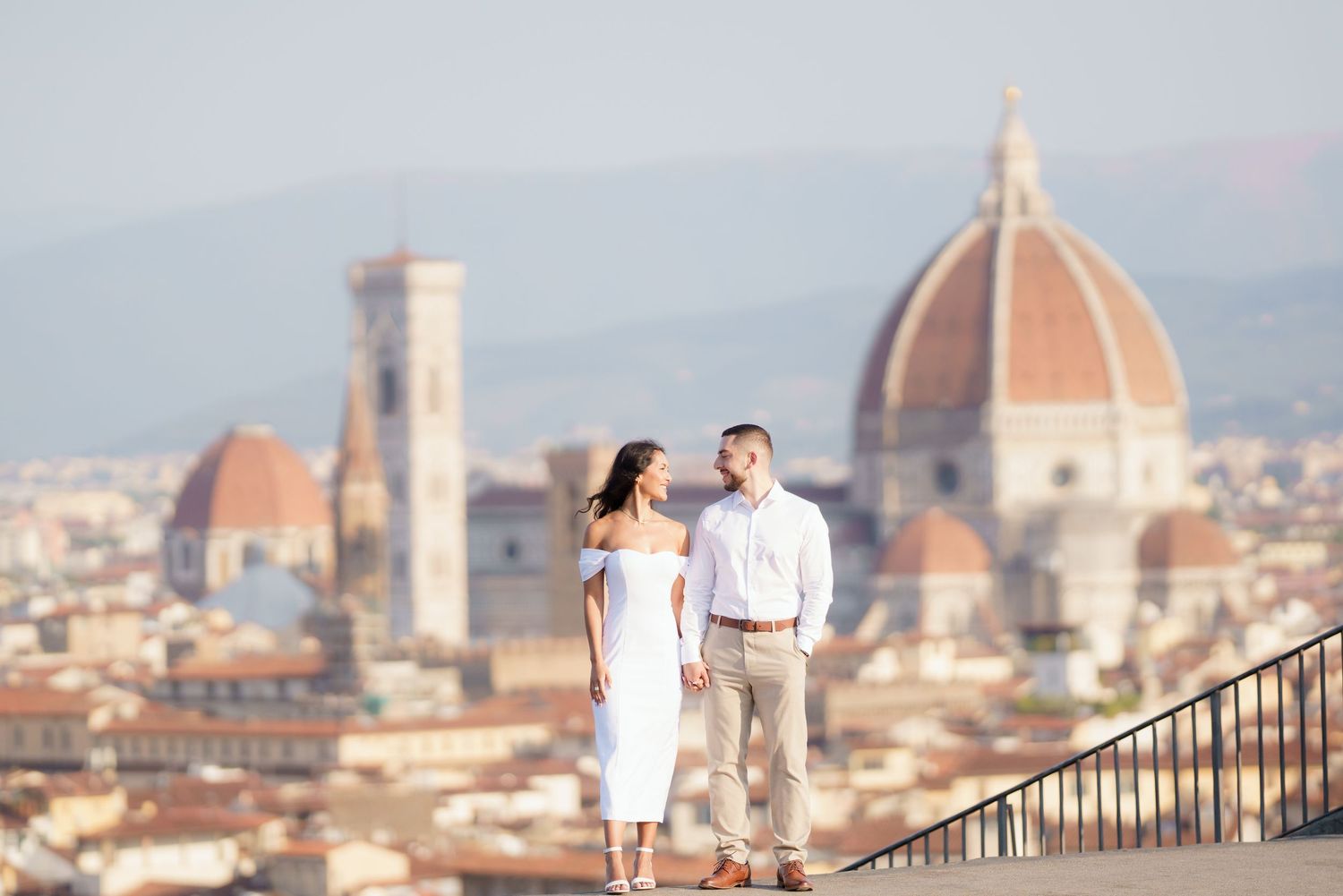 A couple in white outfits poses on a scenic overlook with Florence's iconic Duomo cathedral visible in the background.