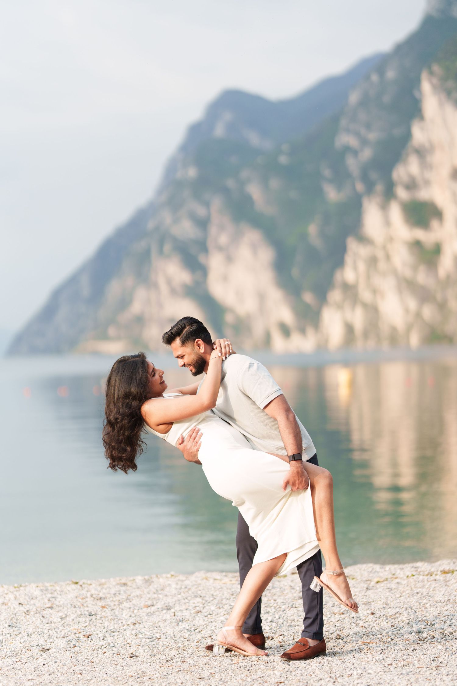 Romantic couple shares tender moment on pebbled beach with dramatic mountain cliffs and lake in background.
