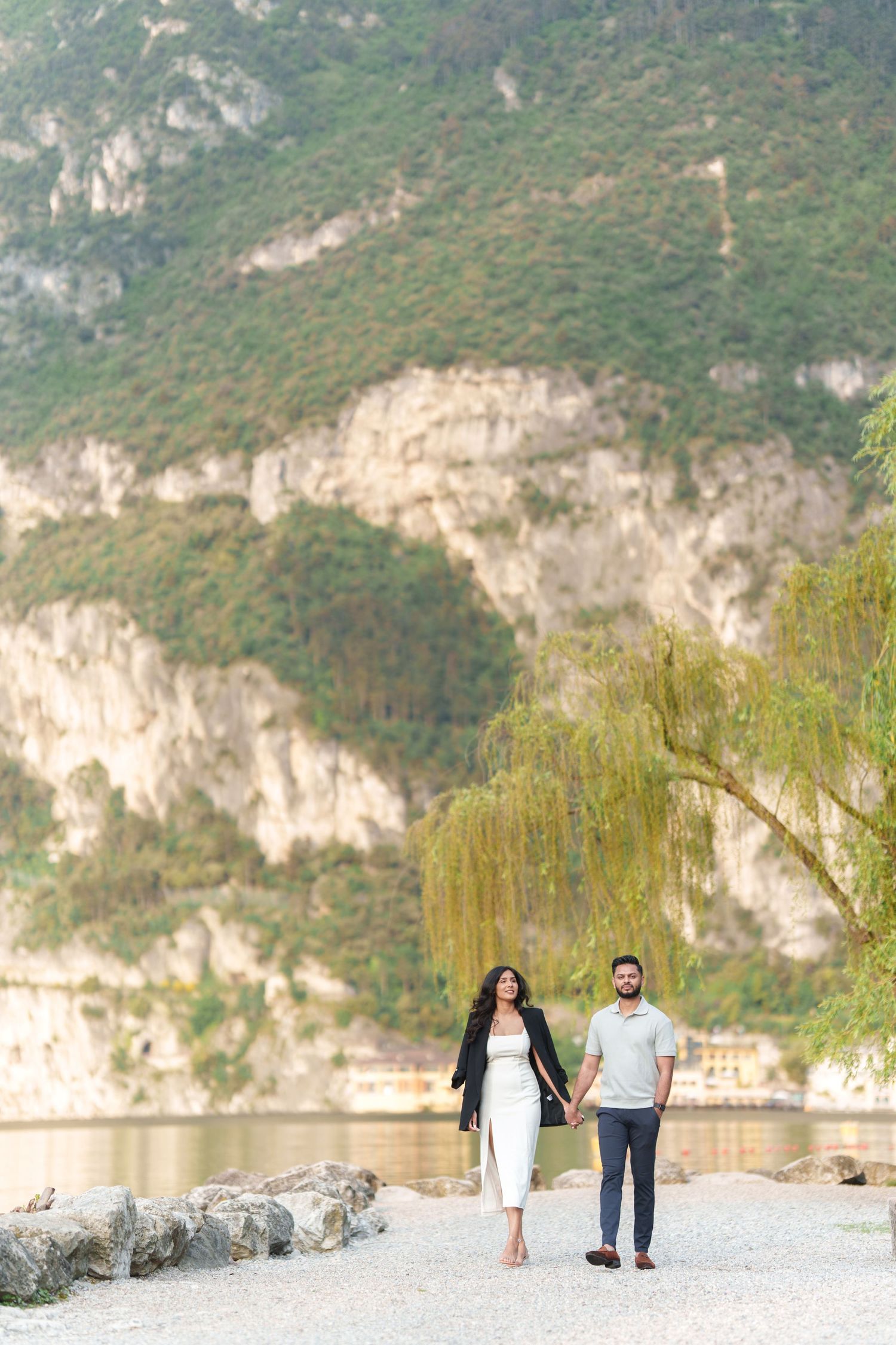 A couple walks along a scenic lakeside path with dramatic limestone cliffs and mountains in the background.