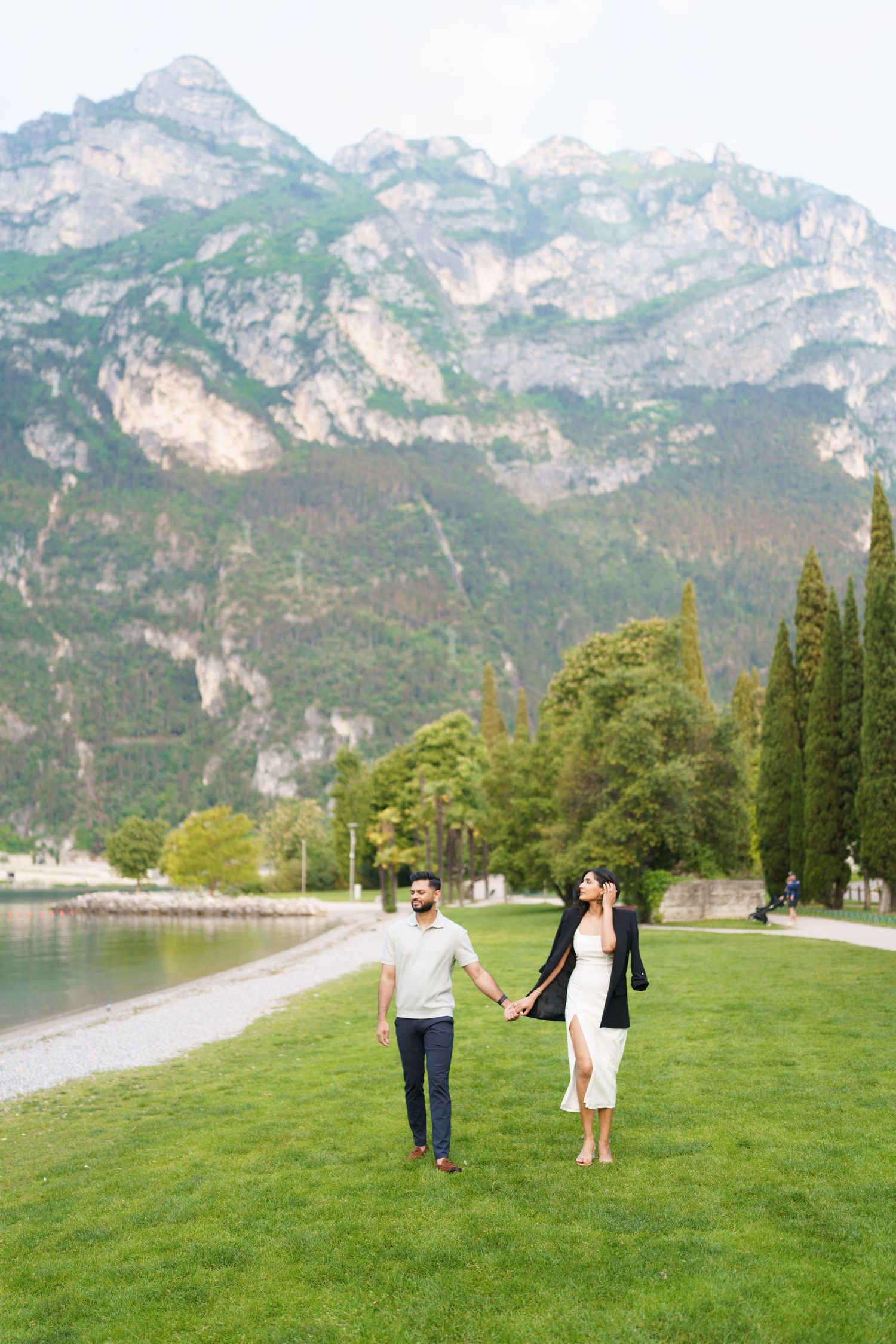 Couple walking hand in hand on green lawn by lake with dramatic mountain peaks rising in background at Lake Garda, Italy.