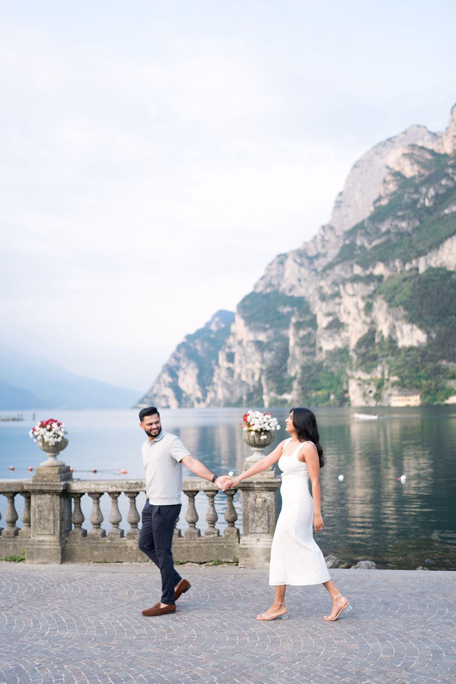 Couple playfully poses near stone balustrade overlooking scenic Lake Garda with mountain backdrop in Italy.