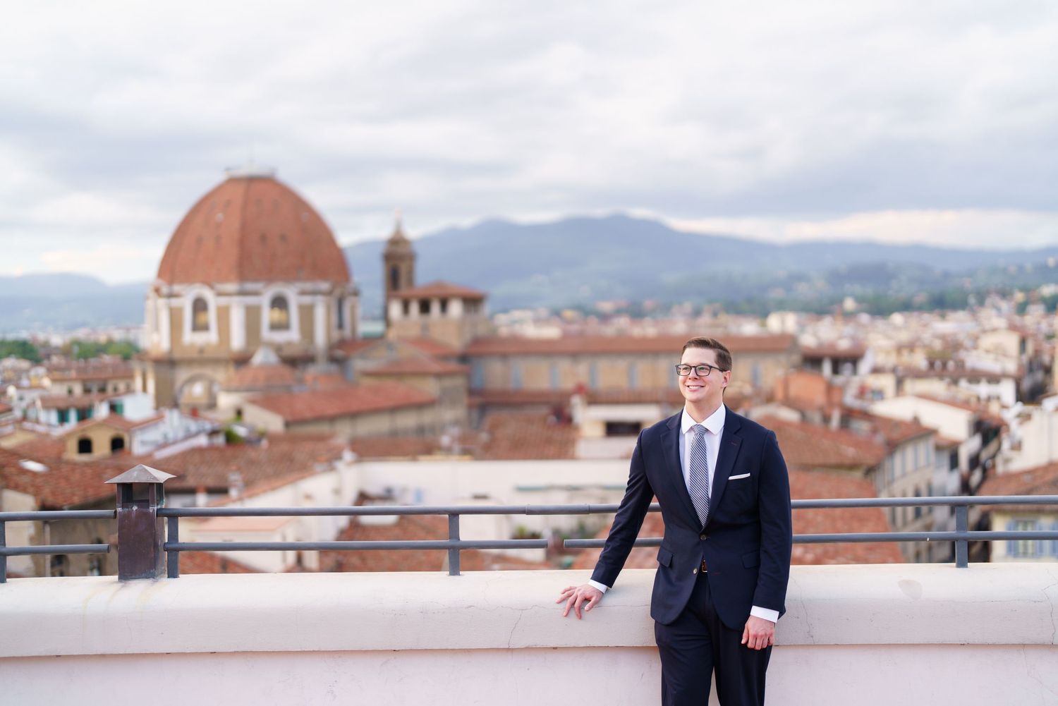 A person in a navy suit holds red roses on a rooftop terrace overlooking Florence, Italy with the Duomo visible.