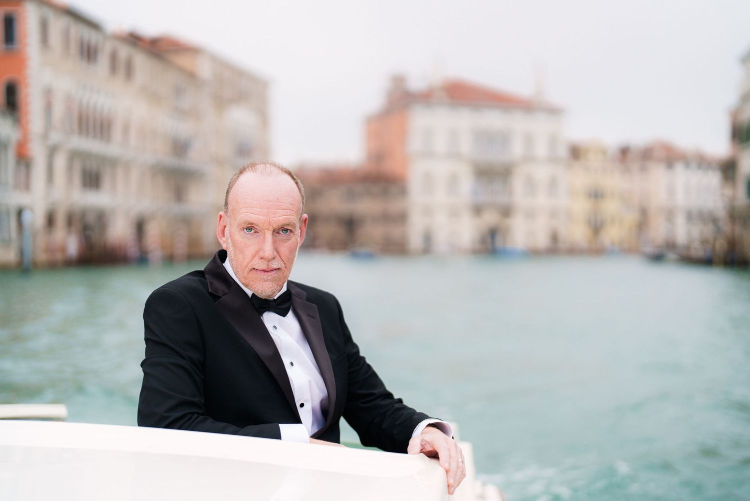 Person in black tuxedo sitting on boat along Venice canal with historic buildings in background.